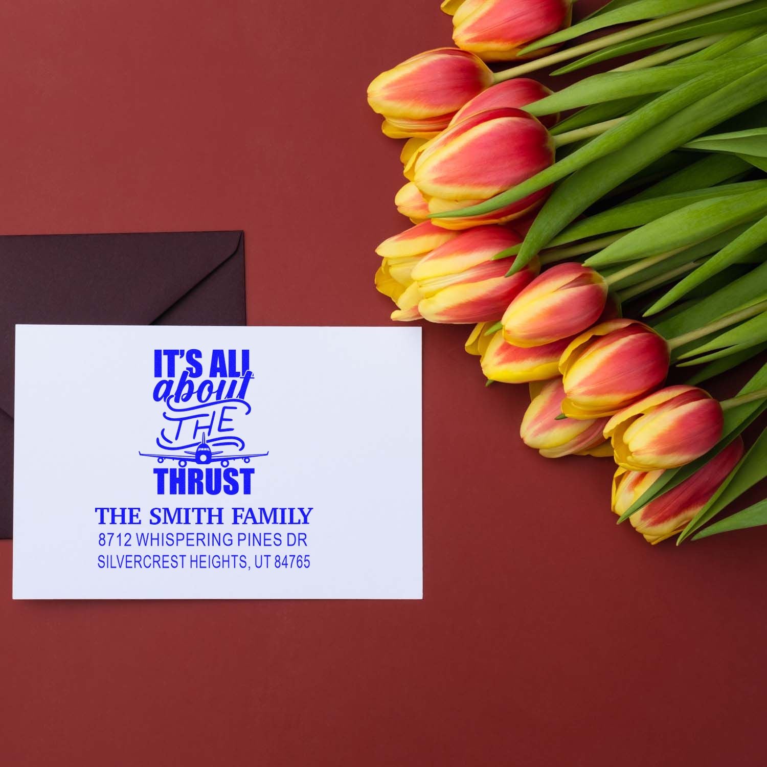 Custom family address stamp with tulips and envelope on burgundy background, featuring blue Its all about the thrust airplane design.