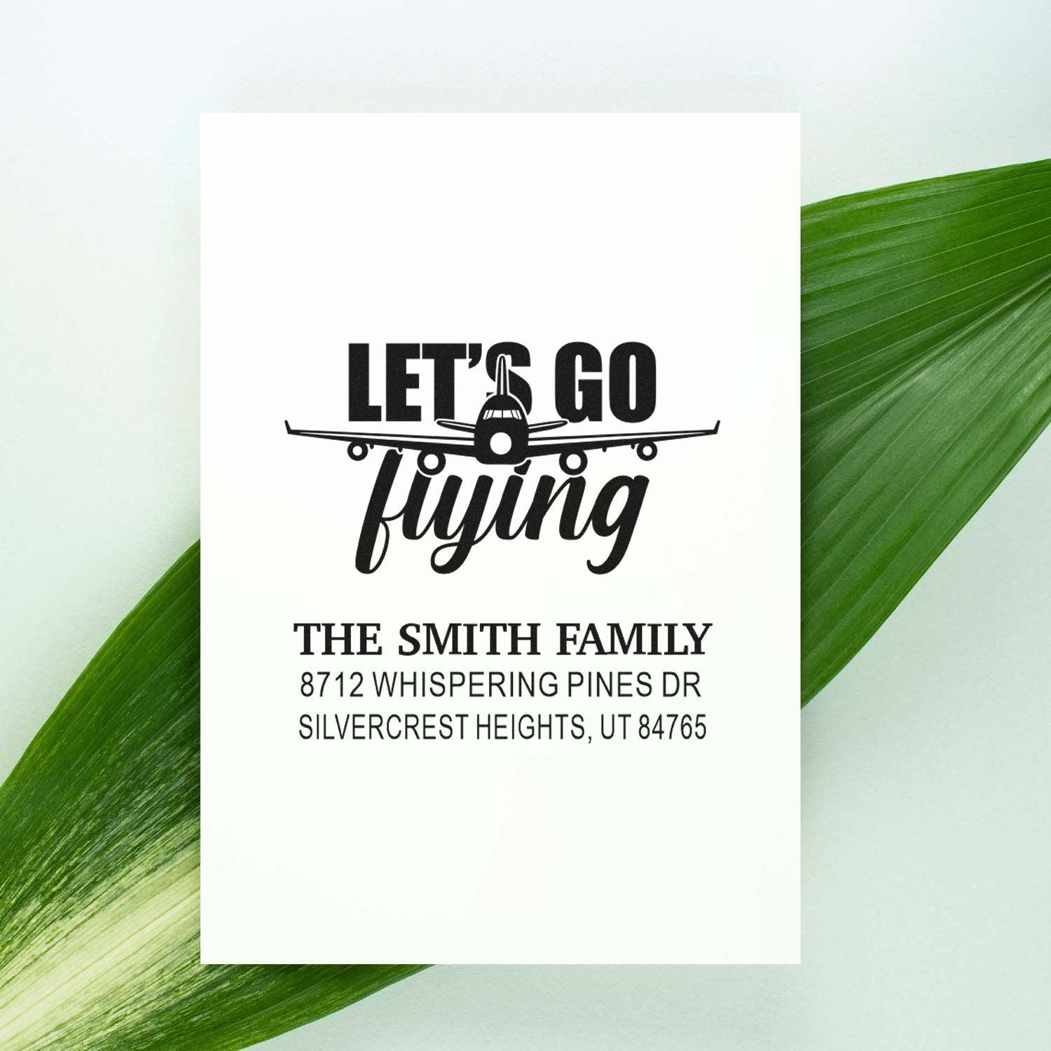 Personalized return address stamp with airplane Lets Go Flying design for The Smith Family, shown on a white card over green leaves.