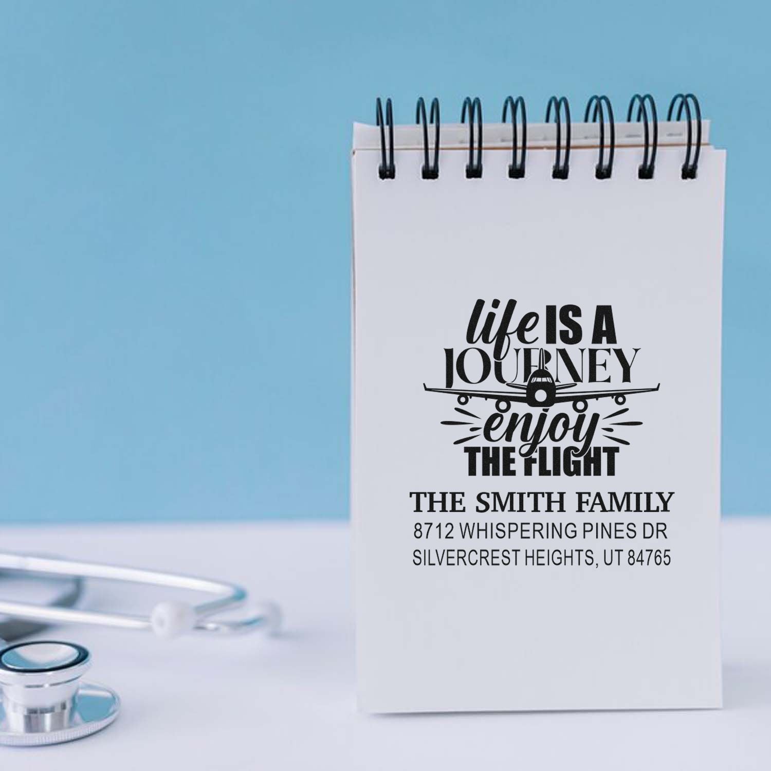 Personalized custom address stamp on a spiral notepad, featuring an airplane graphic and Life is a journey, enjoy the flight on a desk.