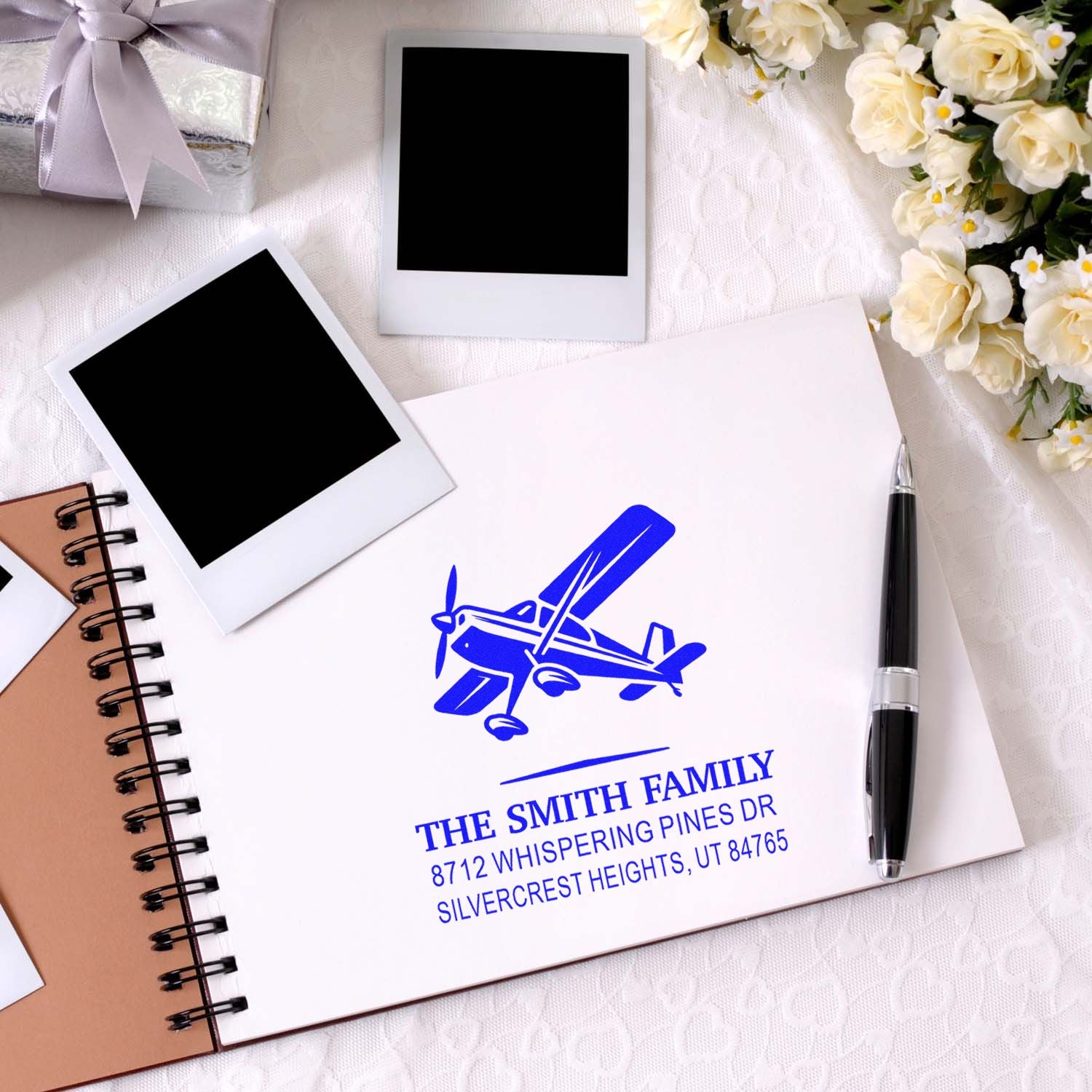 Personalized blue family return address stamp with airplane design on a wedding-themed desk beside Polaroids, pen, notebook, and cream flowers.