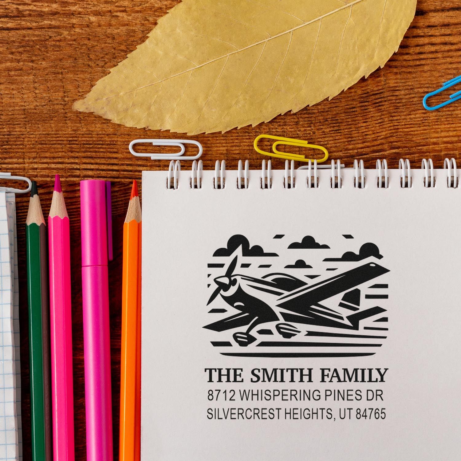 Custom family address stamp with airplane design on a spiral notebook, beside colorful pencils, paper clips, and a leaf on a wooden desk.