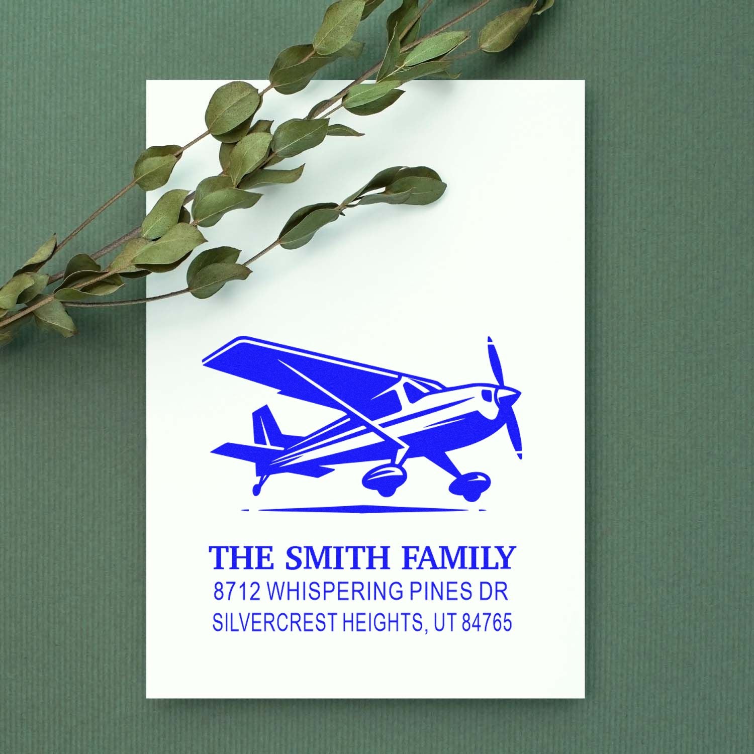 Custom blue airplane address stamp on a white card, styled with eucalyptus sprig on a green textured background.