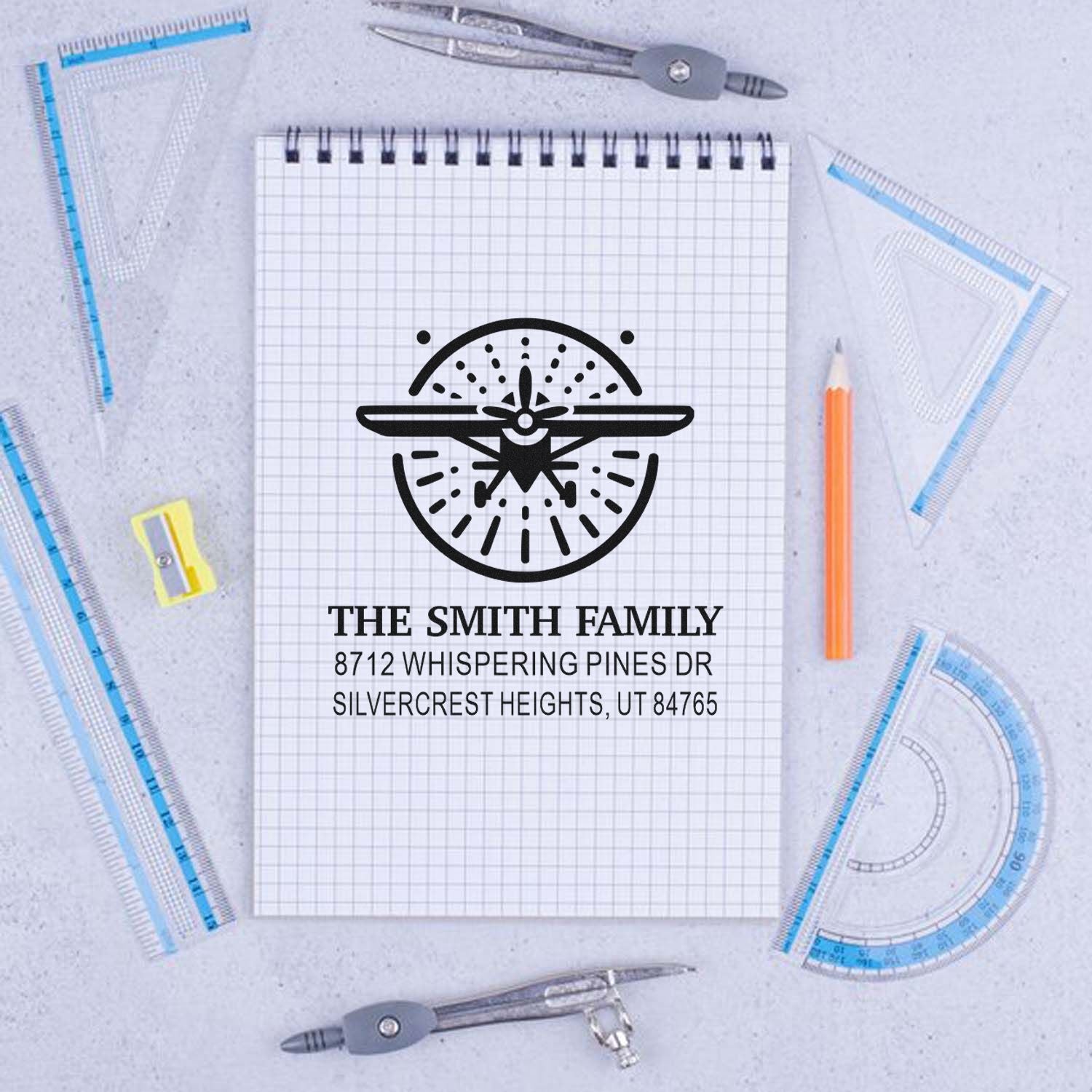 Custom airplane family address stamp on graph paper notebook, surrounded by rulers, compass tools and pencil in a stationery flat lay.