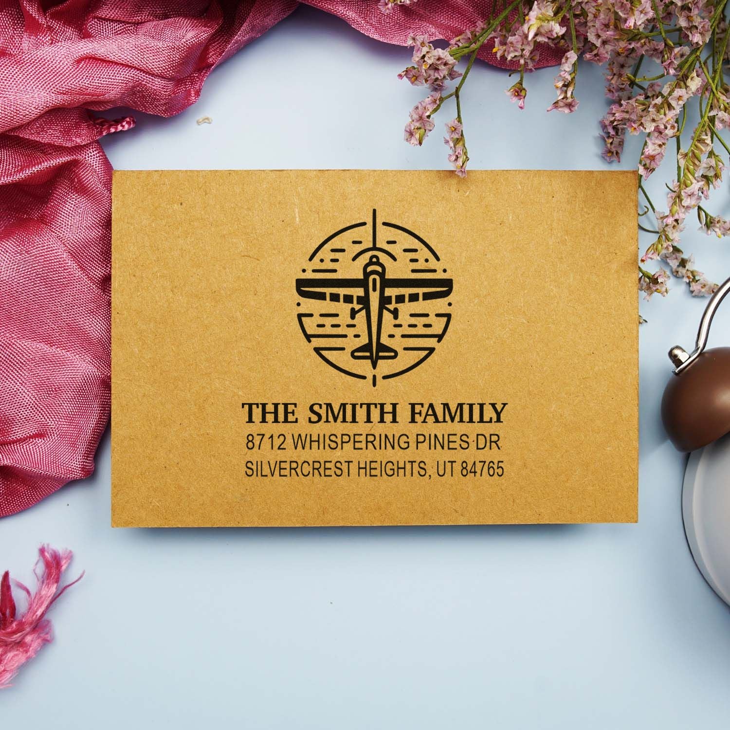 Personalized address stamp with airplane logo for The Smith Family, styled on kraft paper with pink fabric and flowers on a light blue flat lay background.