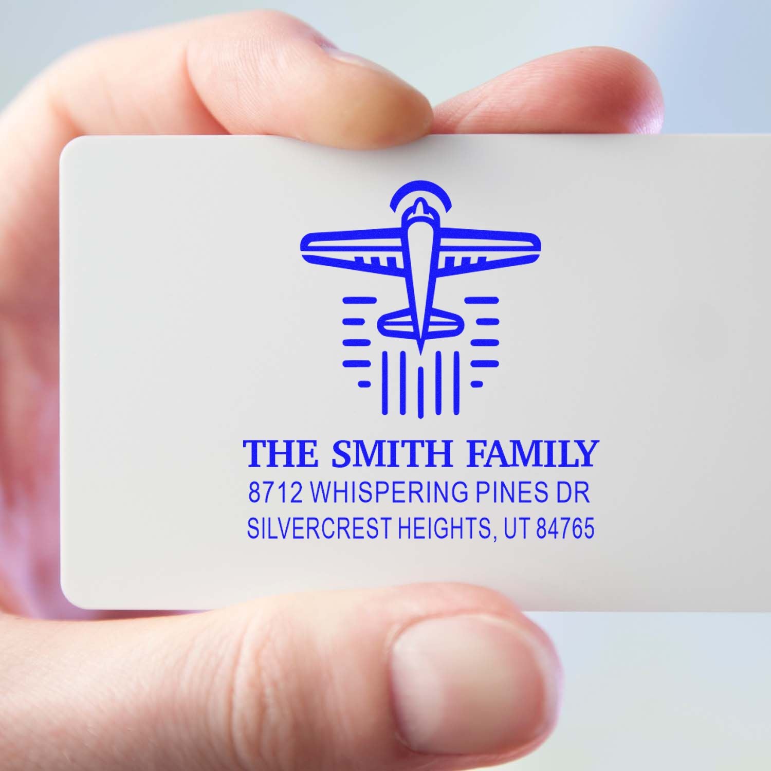 Hand holding a custom blue family address stamp with airplane logo, showing The Smith Family and Silvercrest Heights, UT on a white card.