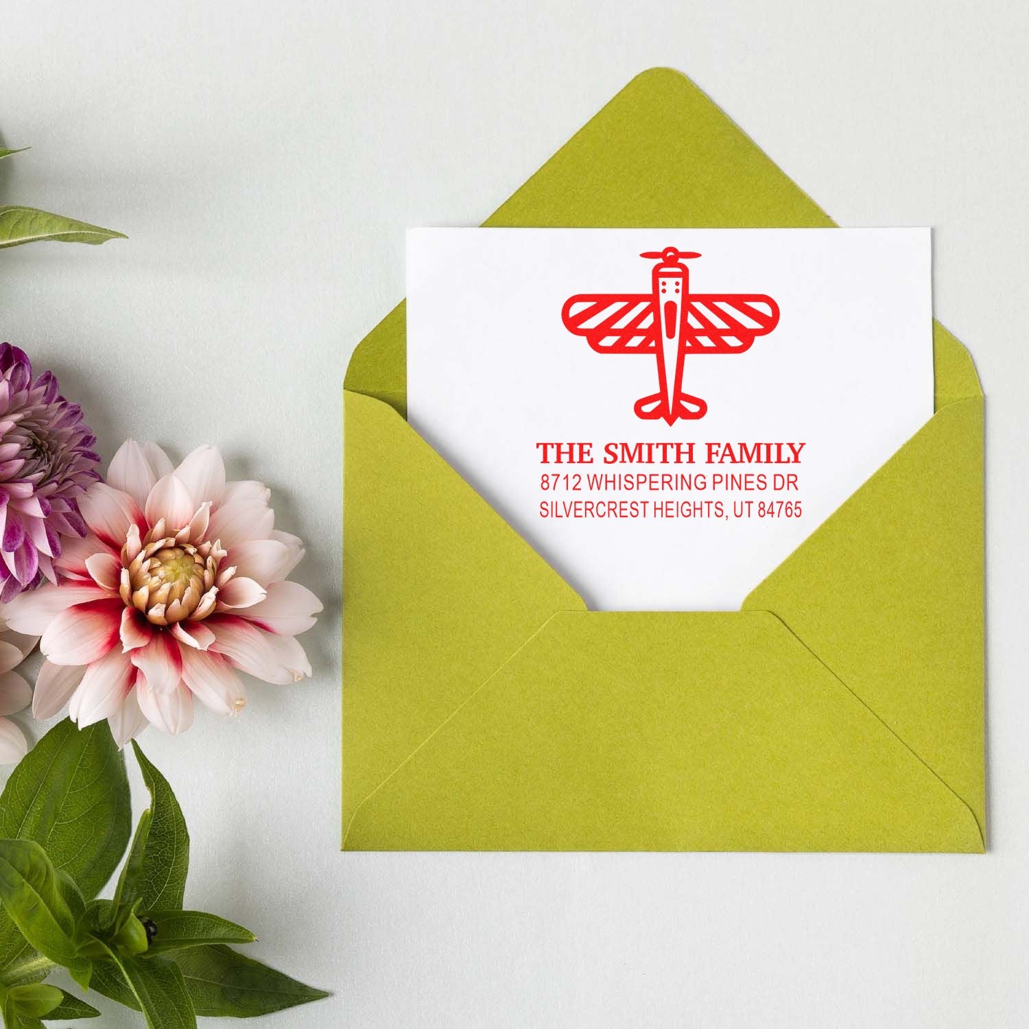 Personalized return address stamp with red airplane design on a white card in a lime green envelope, styled with flowers on a white tabletop.