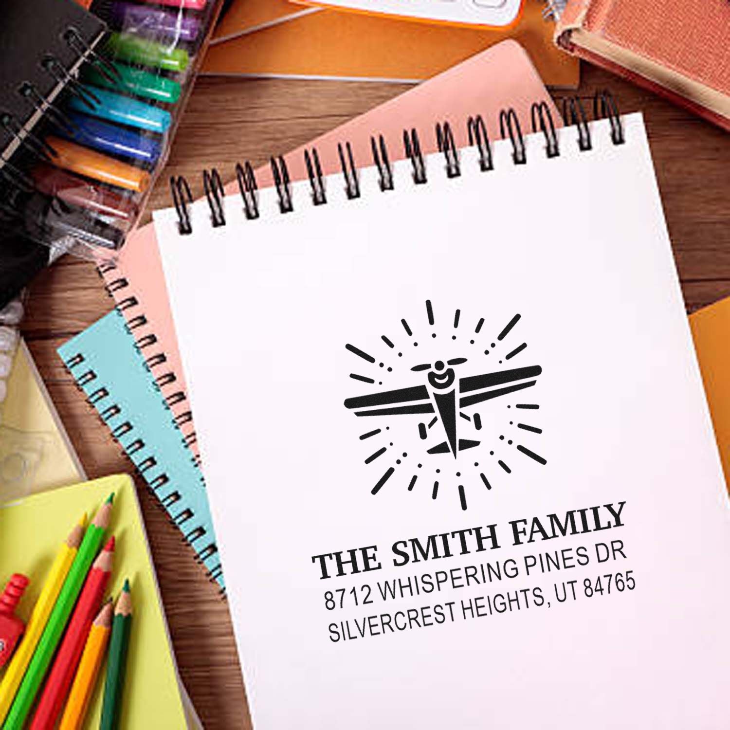 Personalized family return address stamp on spiral notebook, featuring an airplane logo, surrounded by colored pencils and stationery.