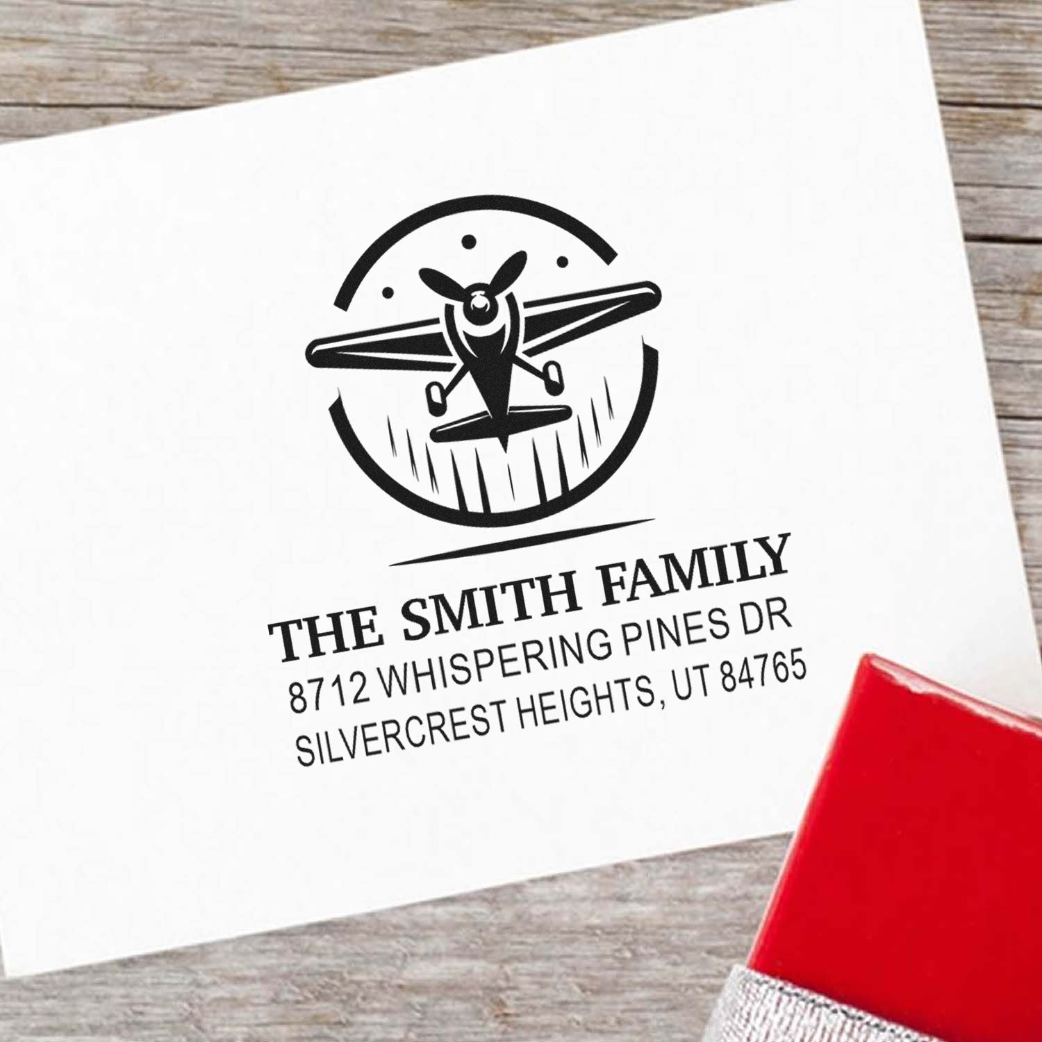 Custom family address stamp with airplane logo on white paper, displayed on a rustic wood table beside a red self-inking stamp handle.