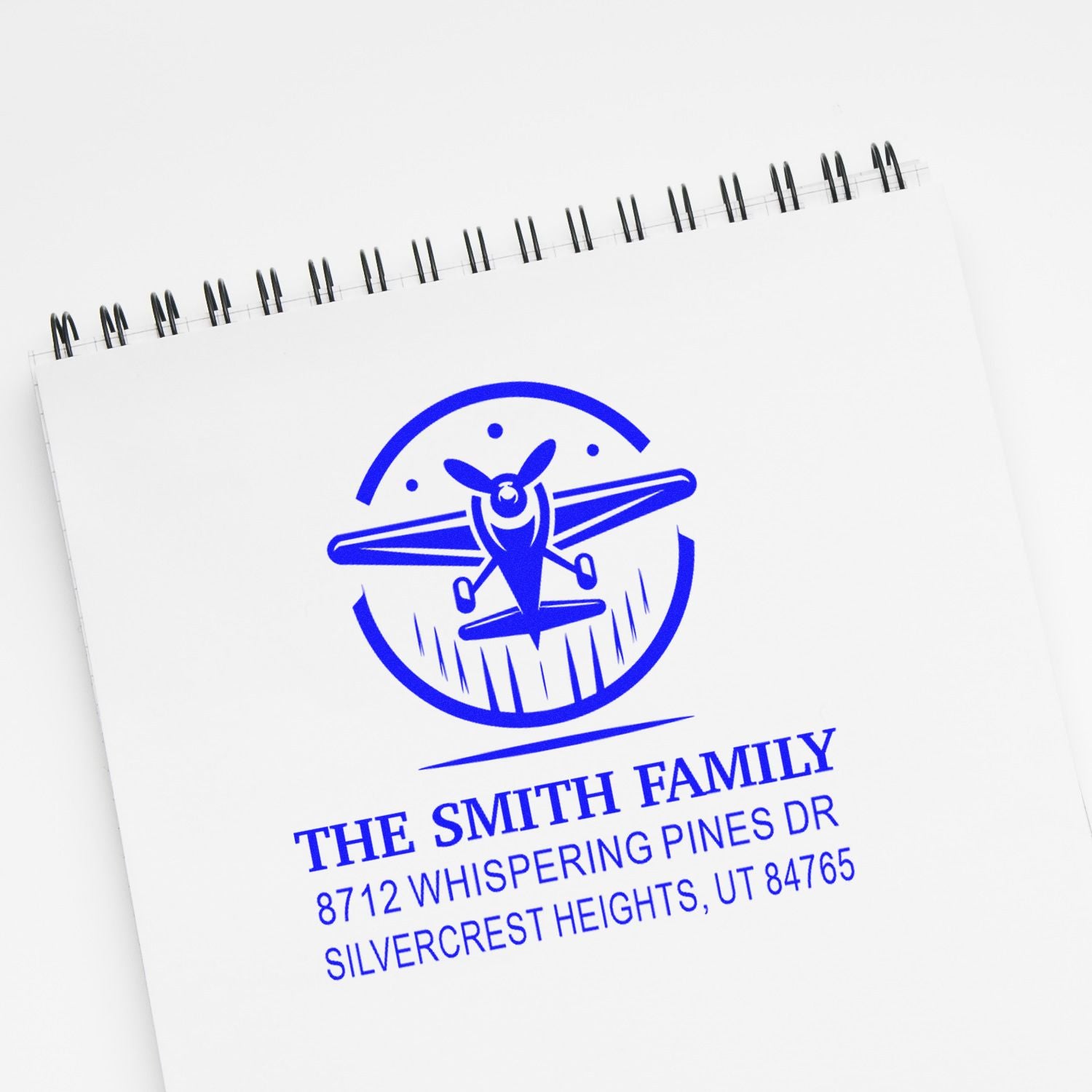Blue custom return address stamp on spiral notebook, featuring airplane logo and The Smith Family address in Silvercrest Heights, UT.