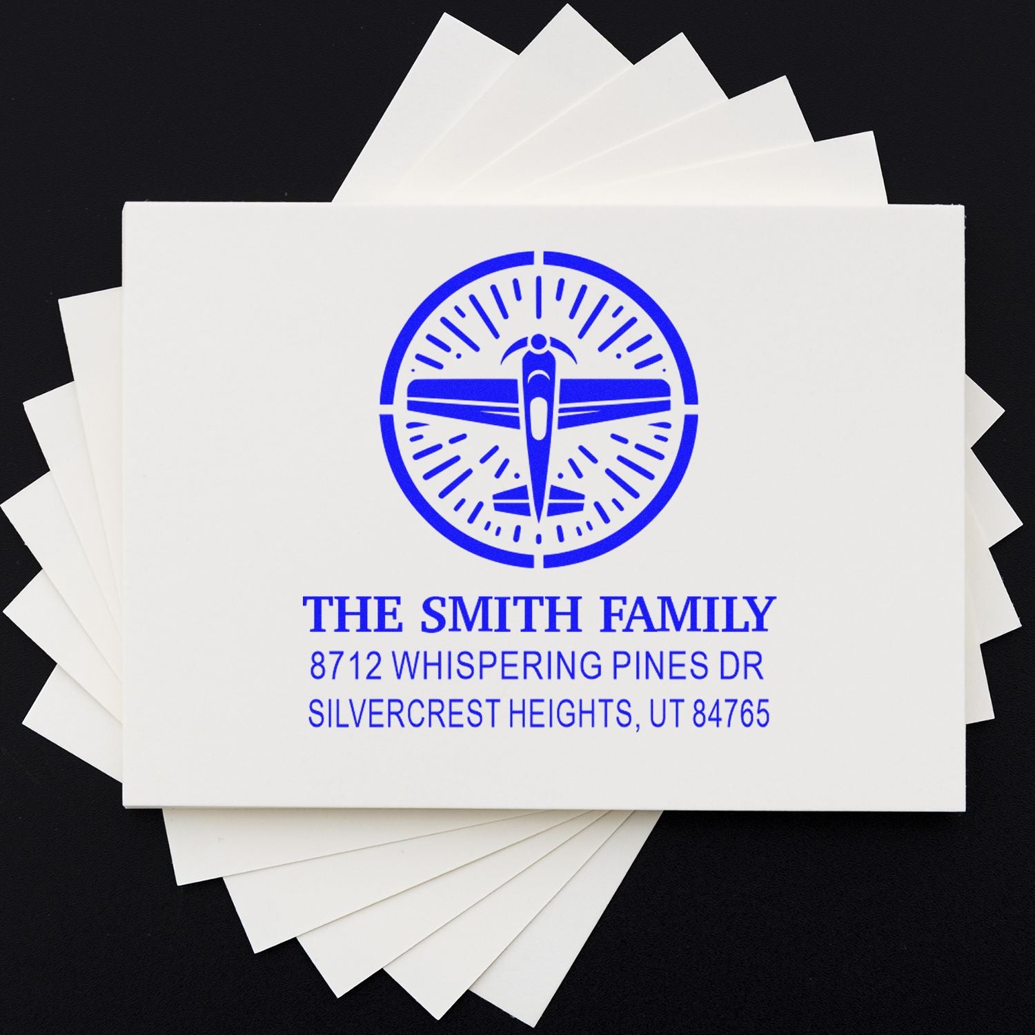 Personalized family return address stamp with blue airplane logo, stacked on white sample cards against a black background.