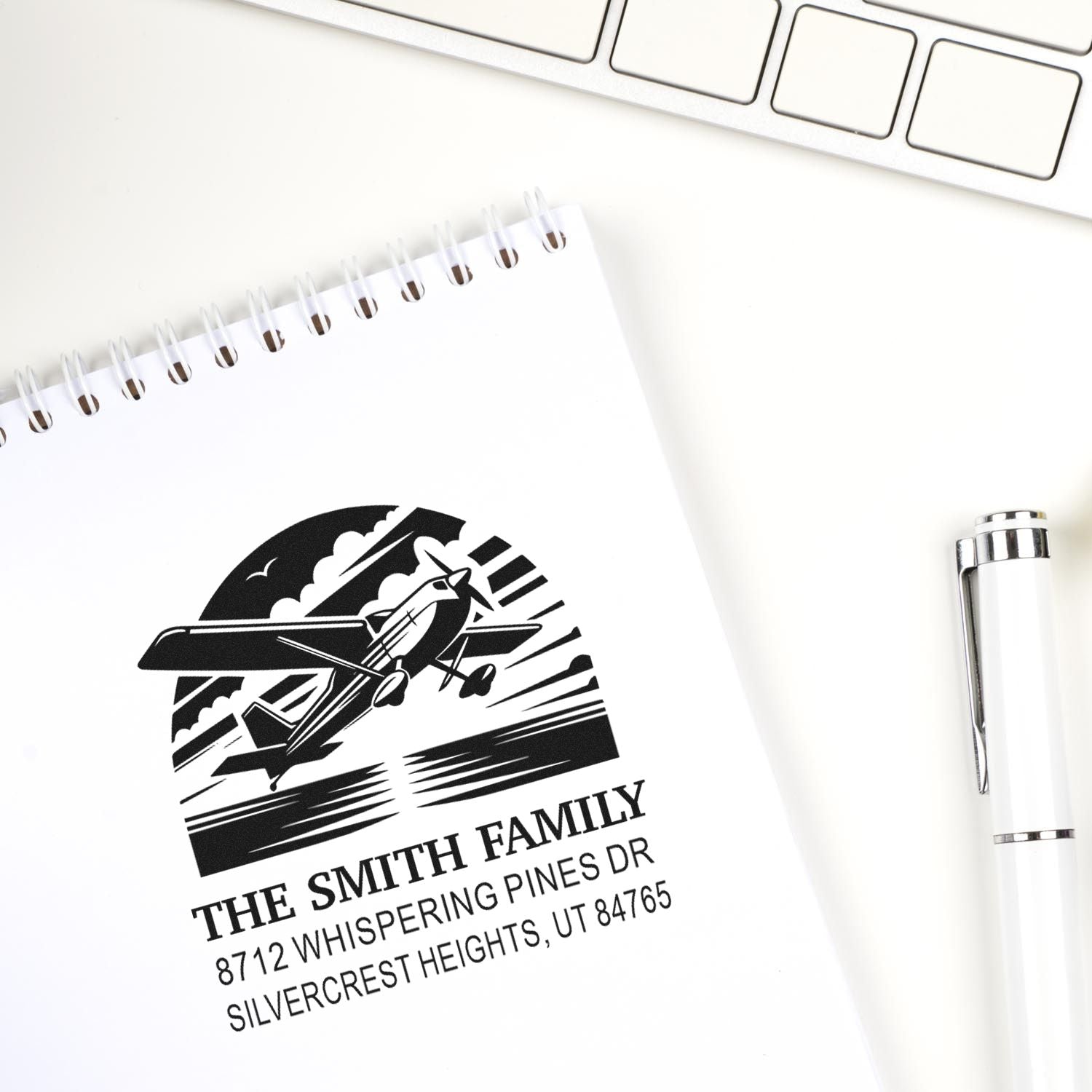 Personalized family address stamp with airplane design on a spiral notebook, shown on a white desk beside a keyboard and pen.
