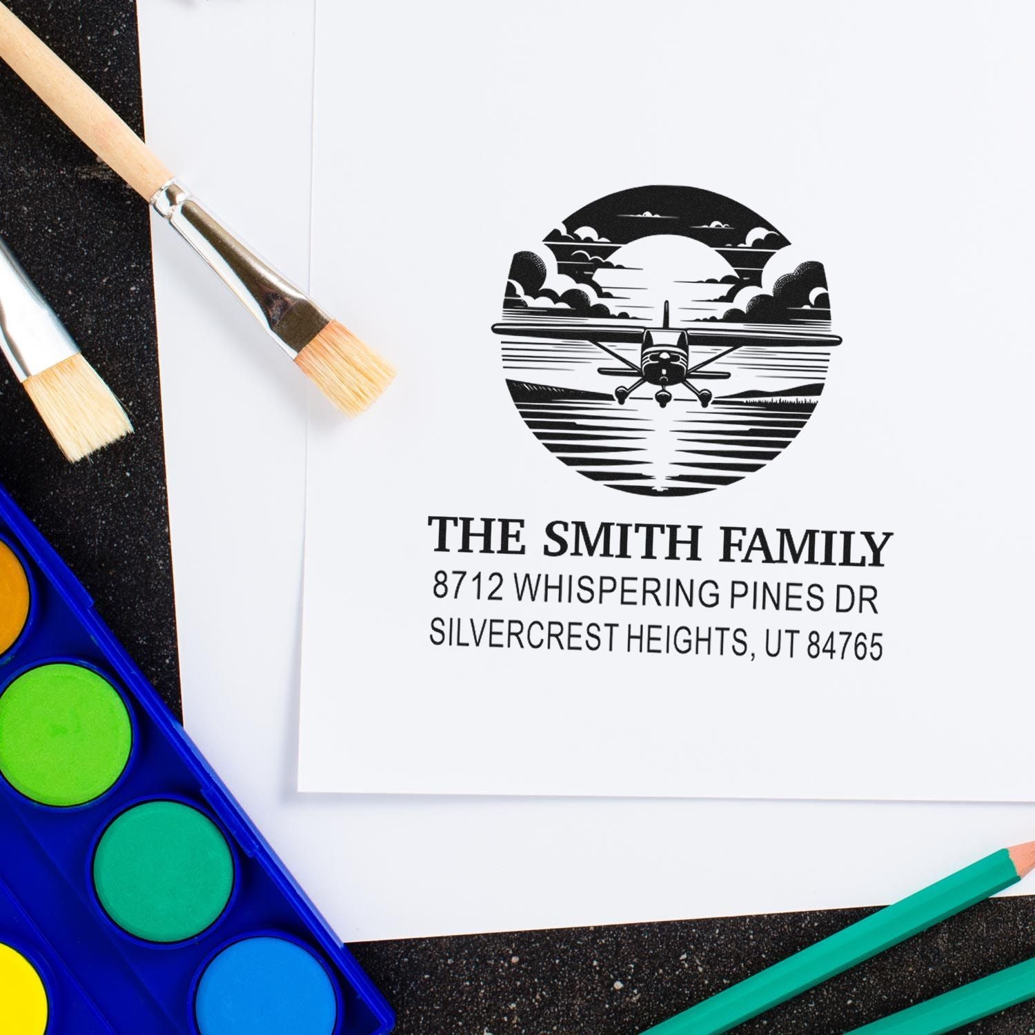 Custom family address stamp with airplane sunset design on paper, surrounded by paintbrushes, watercolor palette, and colored pencils.