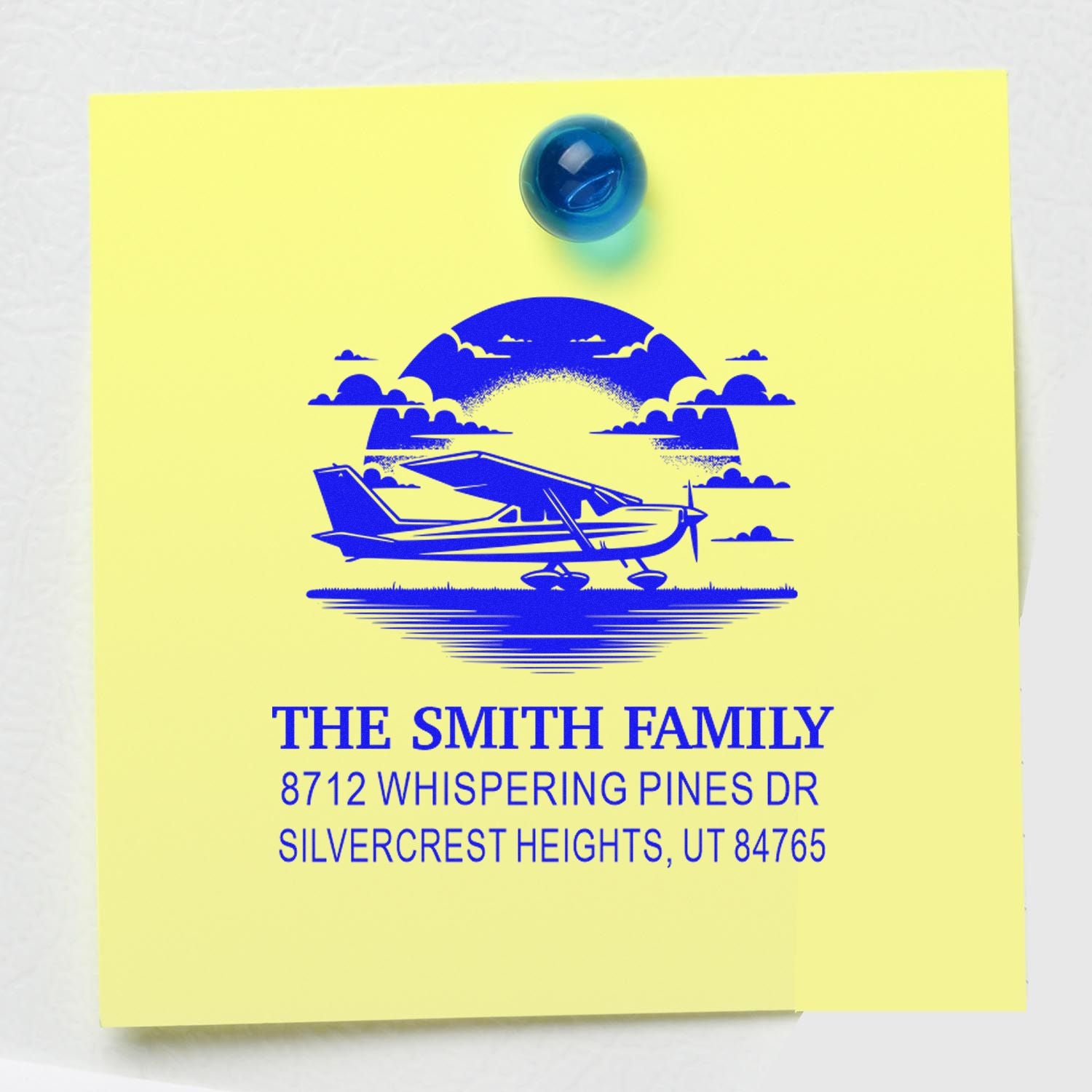 Personalized address stamp with blue airplane graphic and The Smith Family return address on yellow paper, pinned with a blue tack.