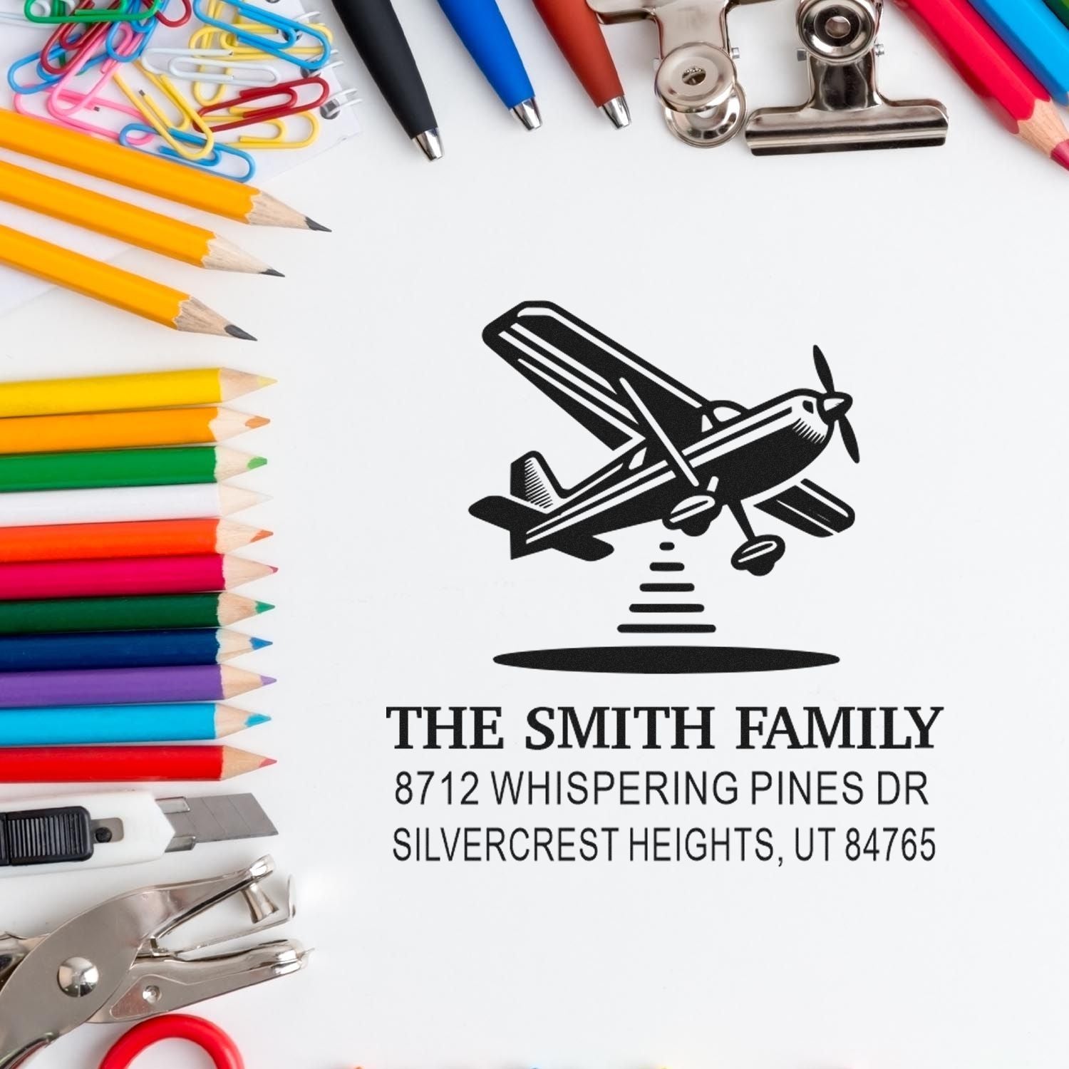 Custom airplane address stamp for The Smith Family, shown with Utah address, surrounded by colorful pencils, paper clips, pens, and office supplies.