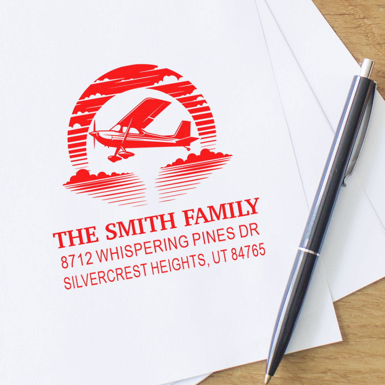 Custom red personalized family address stamp with airplane design on white paper, beside a silver pen on a wooden desk.