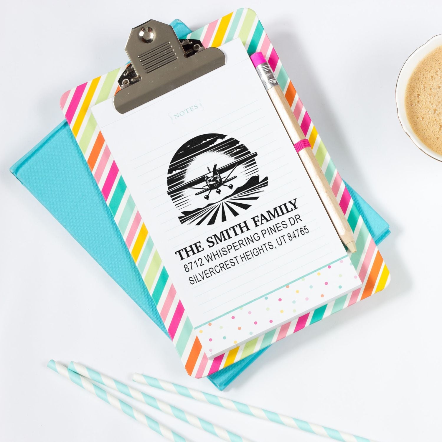 Personalized family address stamp on striped clipboard with notebook, pencil, coffee cup and pastel straws on a white desk.