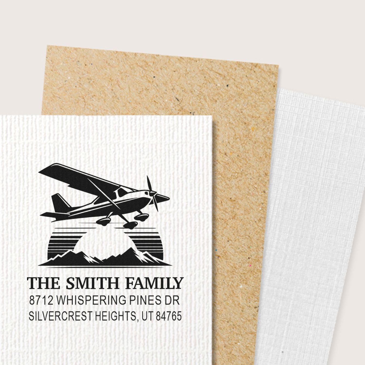 Custom family address stamp with airplane and mountain design on textured cardstock samples, featuring The Smith Family and Silvercrest Heights address.