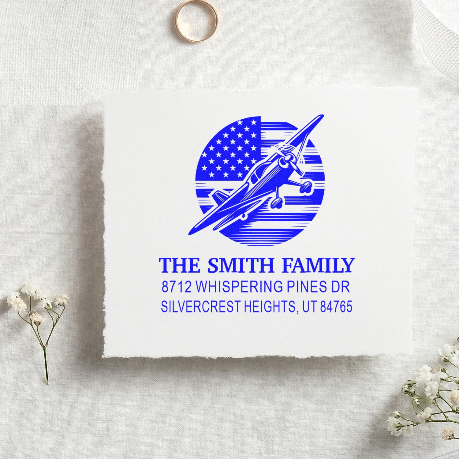 Custom blue personalized address stamp with airplane and American flag design on white paper, styled on a linen backdrop with flowers.