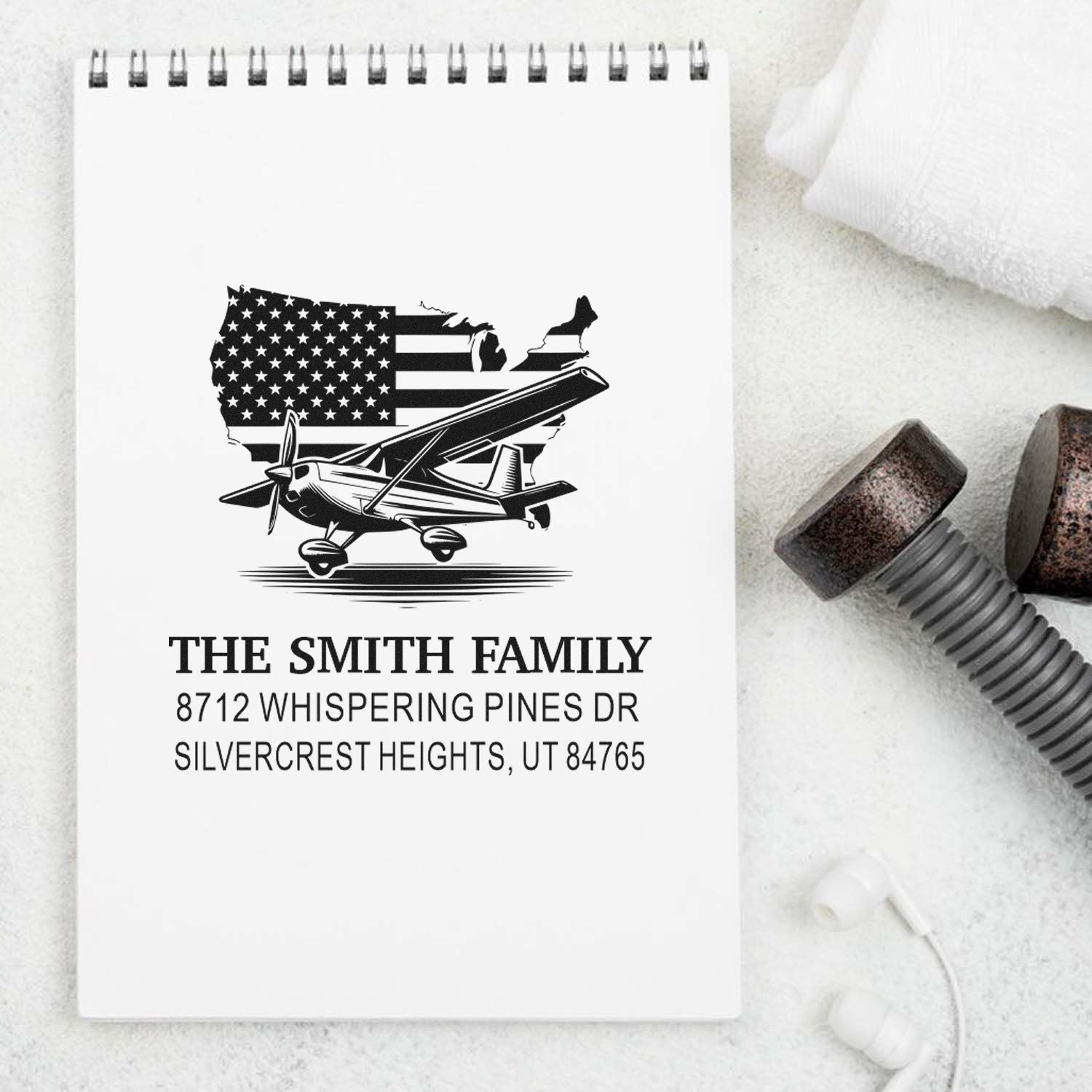 Personalized address stamp with U.S. flag map and airplane design on a notepad, surrounded by stamp handles and earbuds on a white background.