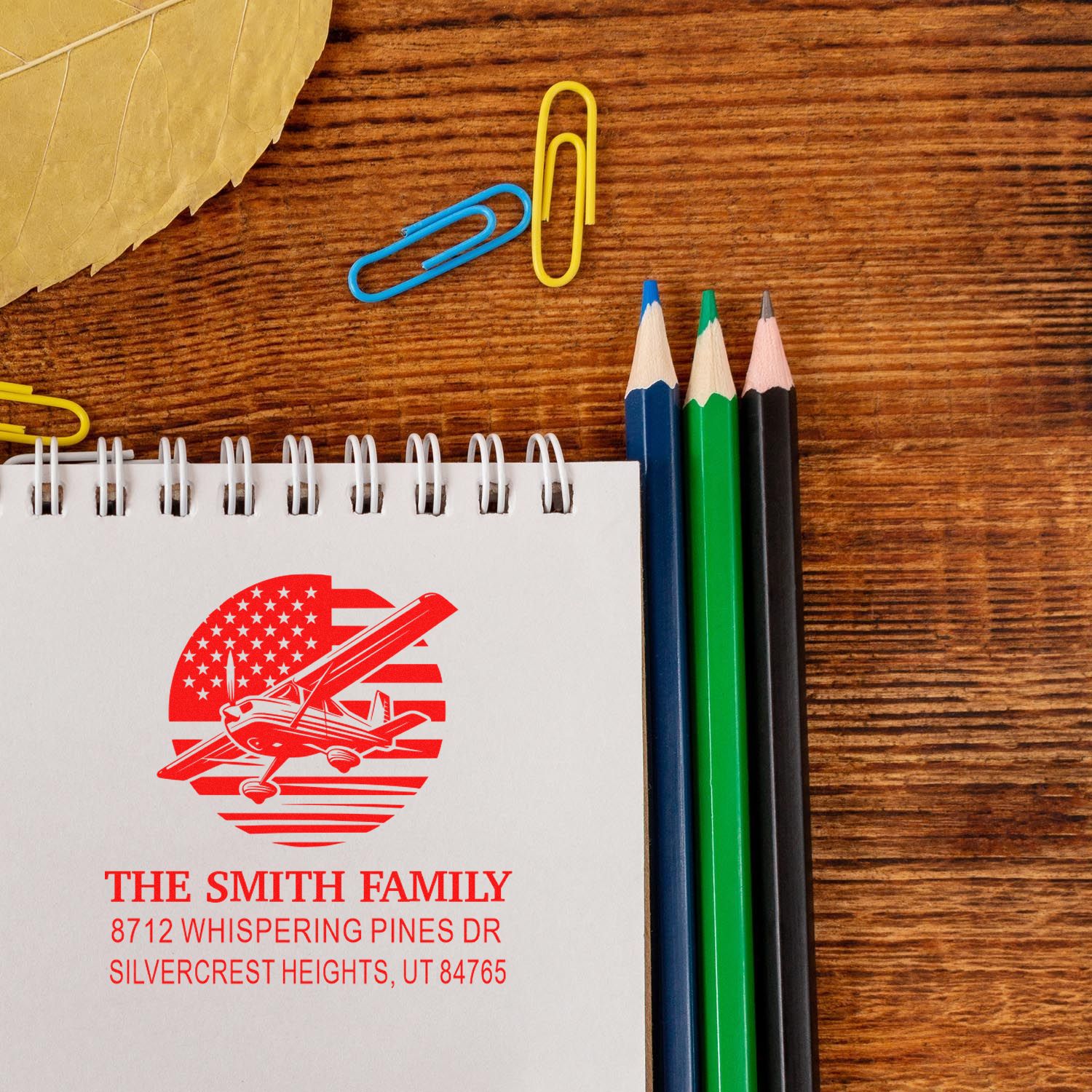 Red personalized family address stamp on a notebook, with airplane logo, beside colored pencils and paper clips on a wooden desk.