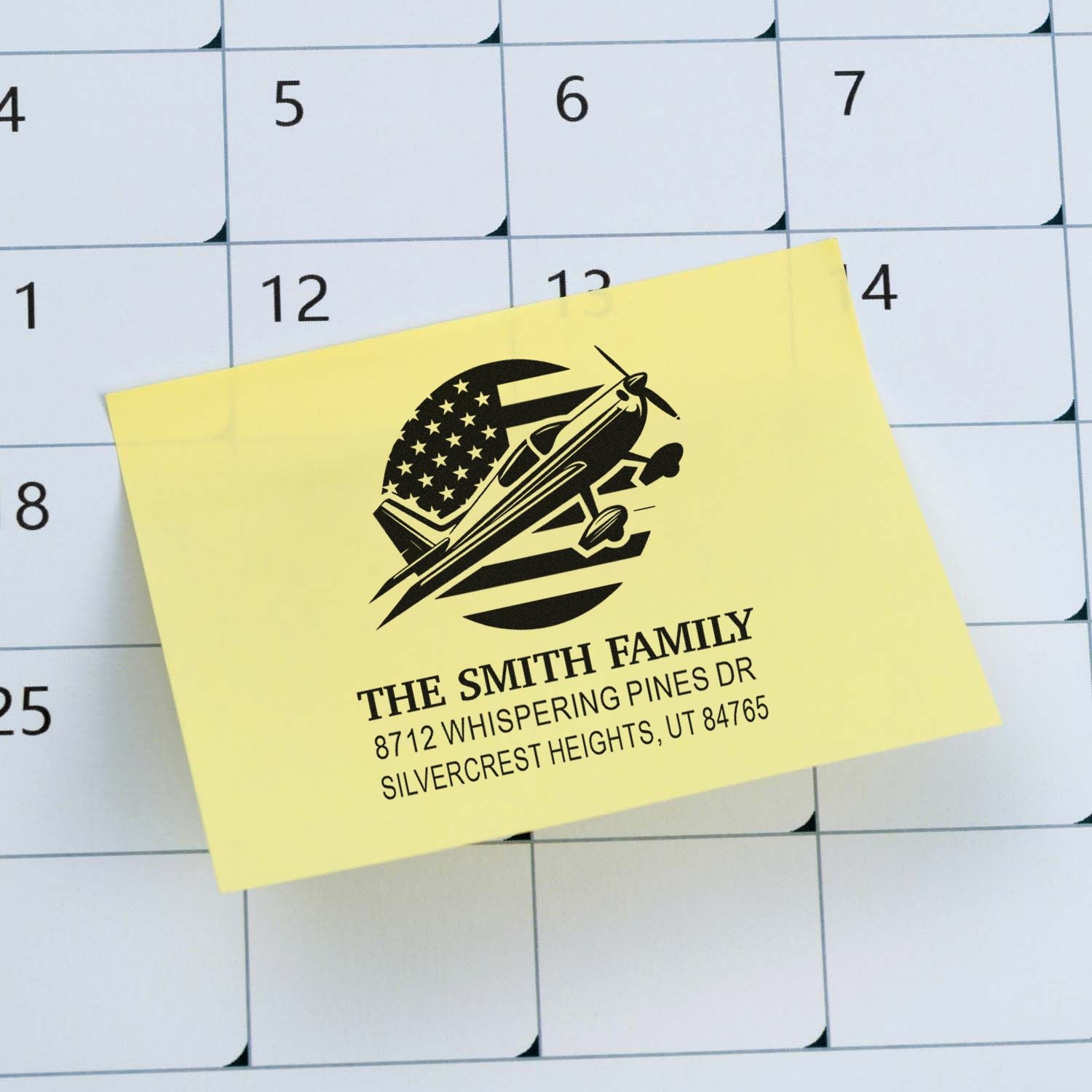 Personalized return address stamp with airplane and U.S. flag design on a yellow note over a wall calendar.