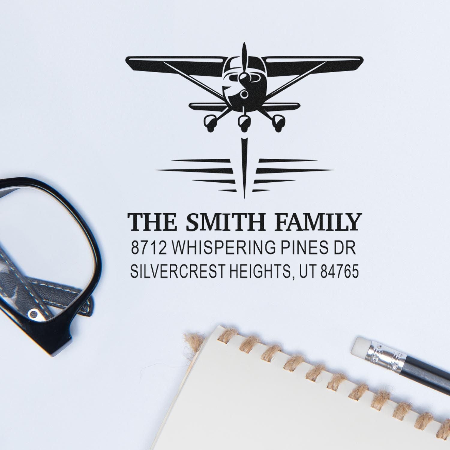 Personalized airplane address stamp with family name and Utah address, shown with stationery, scissors, and pencil on a white desktop.