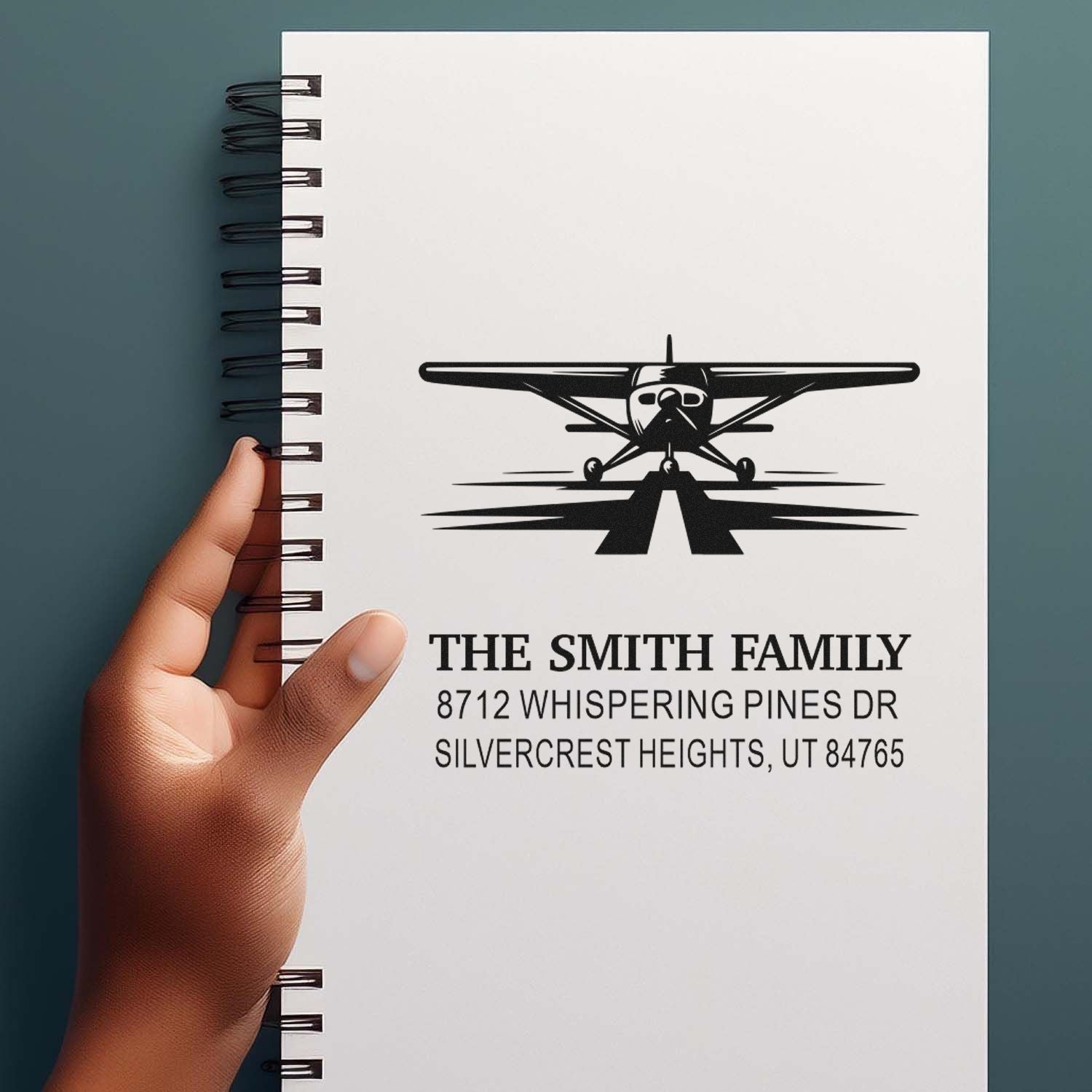 Custom family address stamp with airplane logo on a spiral notebook, showing The Smith Family mailing address, held in hand.