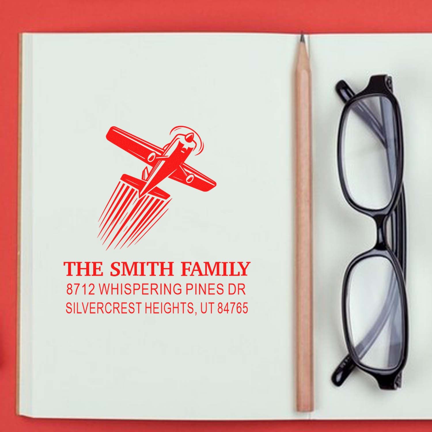 Custom return address stamp with airplane design and Smith family address on an open notebook, beside a pencil and eyeglasses.