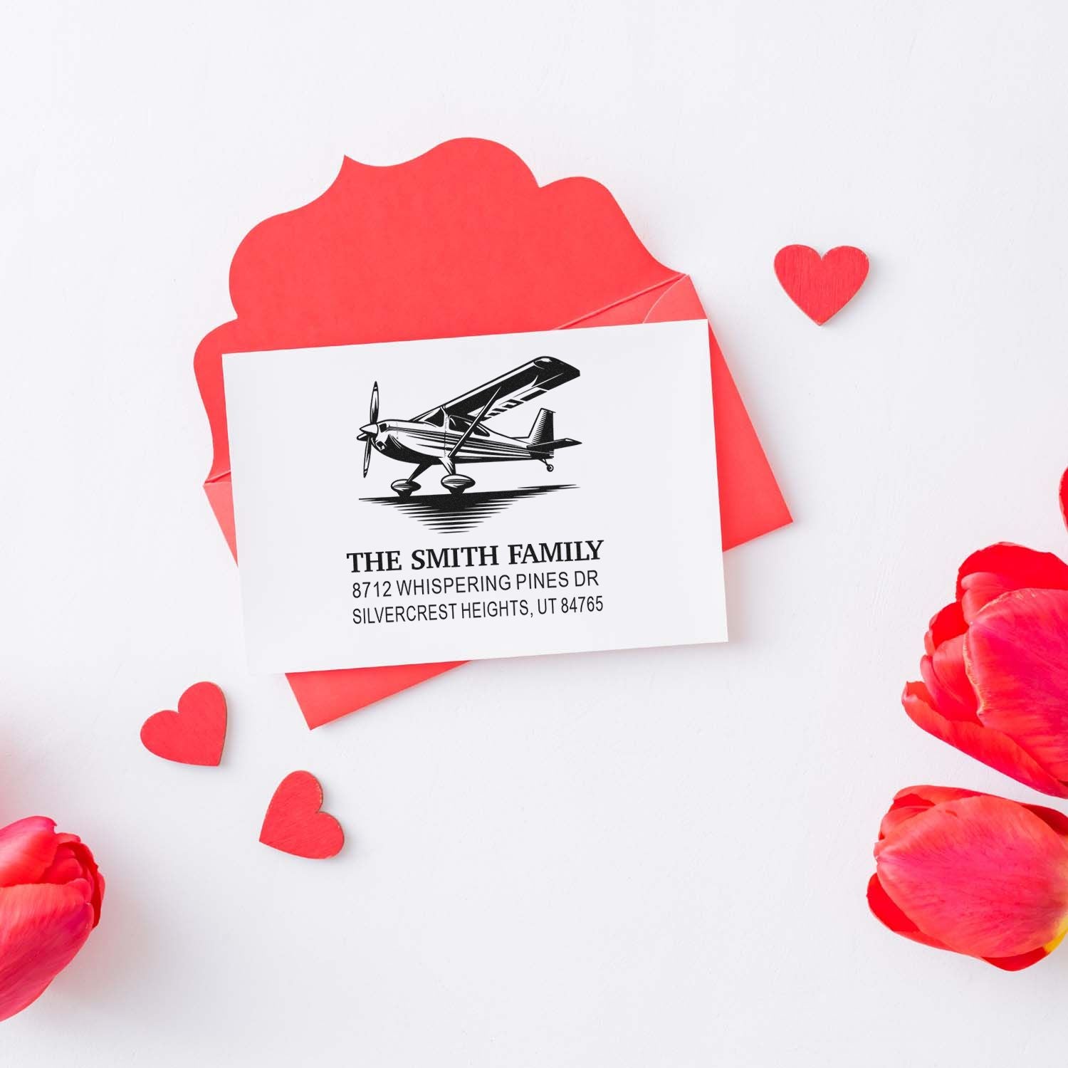 Personalized airplane return address stamp for The Smith Family, displayed with red Valentine hearts, envelope, and floral petals on a white background.