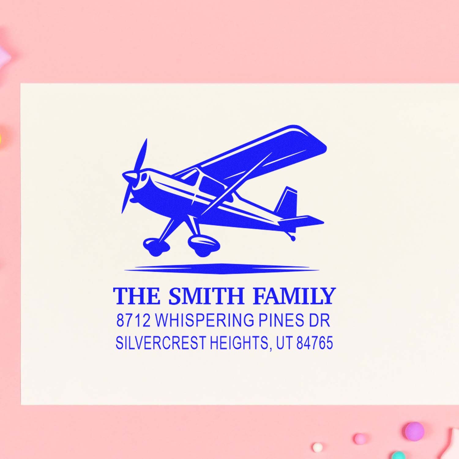Personalized blue airplane address stamp for The Smith Family with Whispering Pines Dr in Silvercrest Heights, on a cream card against a pink background.