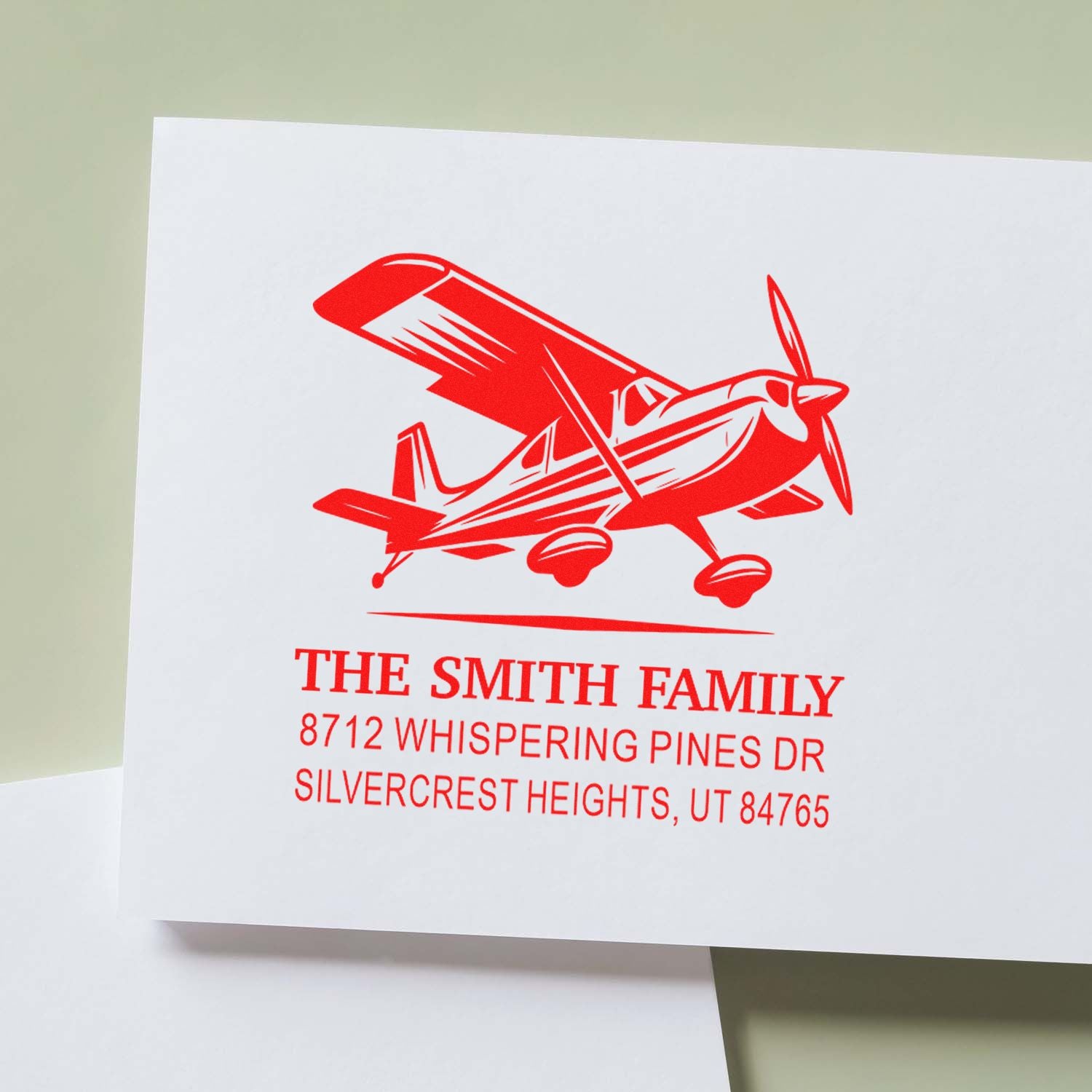Custom red family address stamp with airplane design on white paper, reading The Smith Family, 8712 Whispering Pines Dr, Silvercrest Heights, UT 84765.