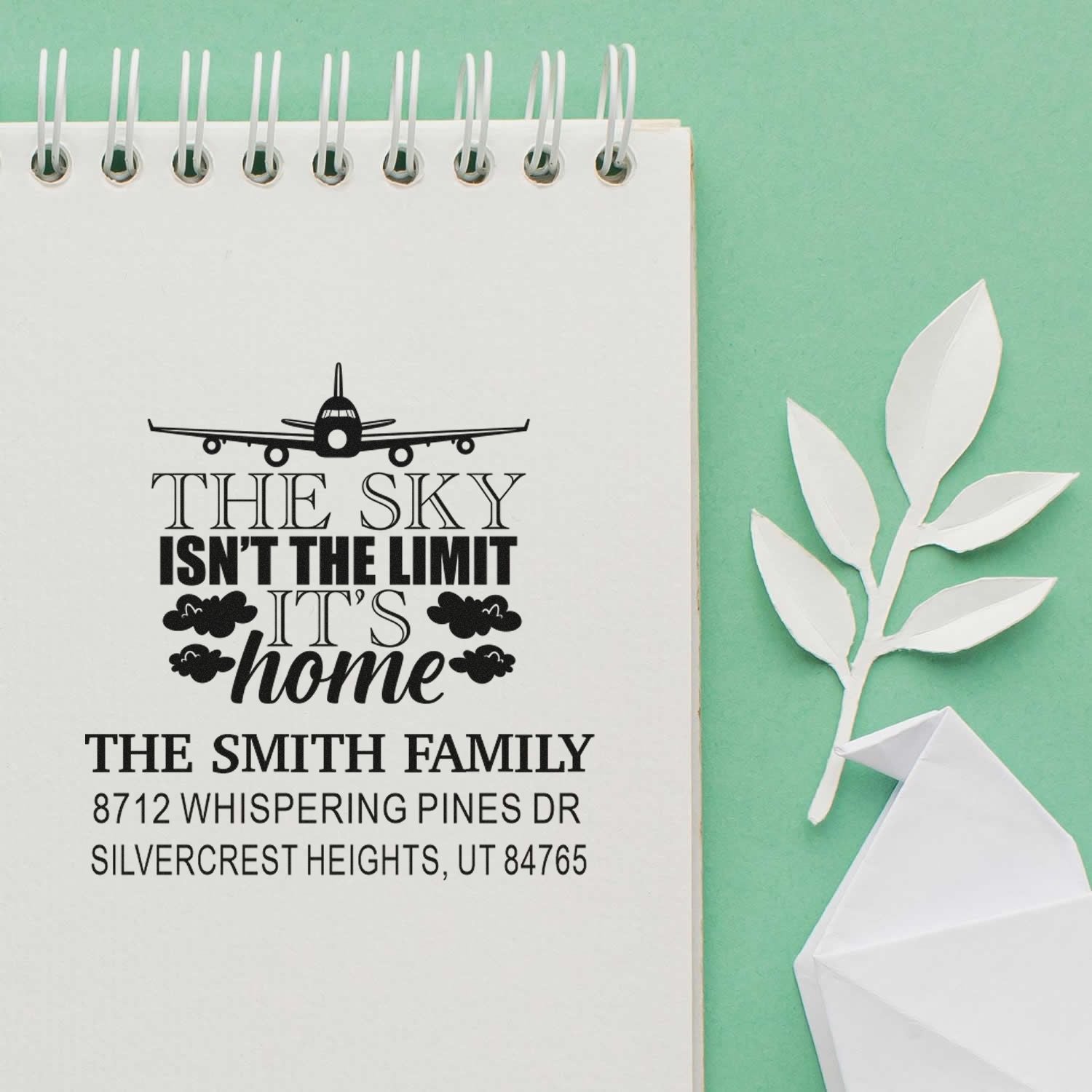 Custom self-inking address stamp with airplane quote and Smith family address on a spiral notebook, styled with paper leaves and an origami bird on mint green.
