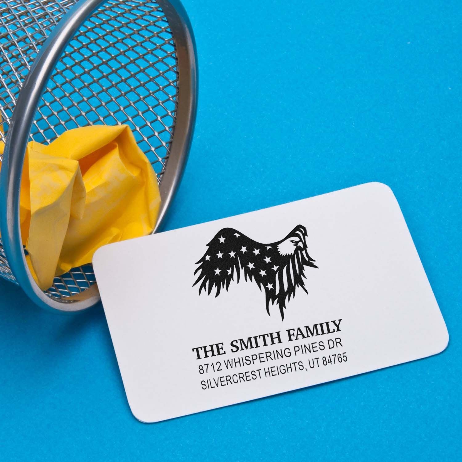 Custom family address stamp with eagle design on a white card beside a metal mesh basket on a blue background.