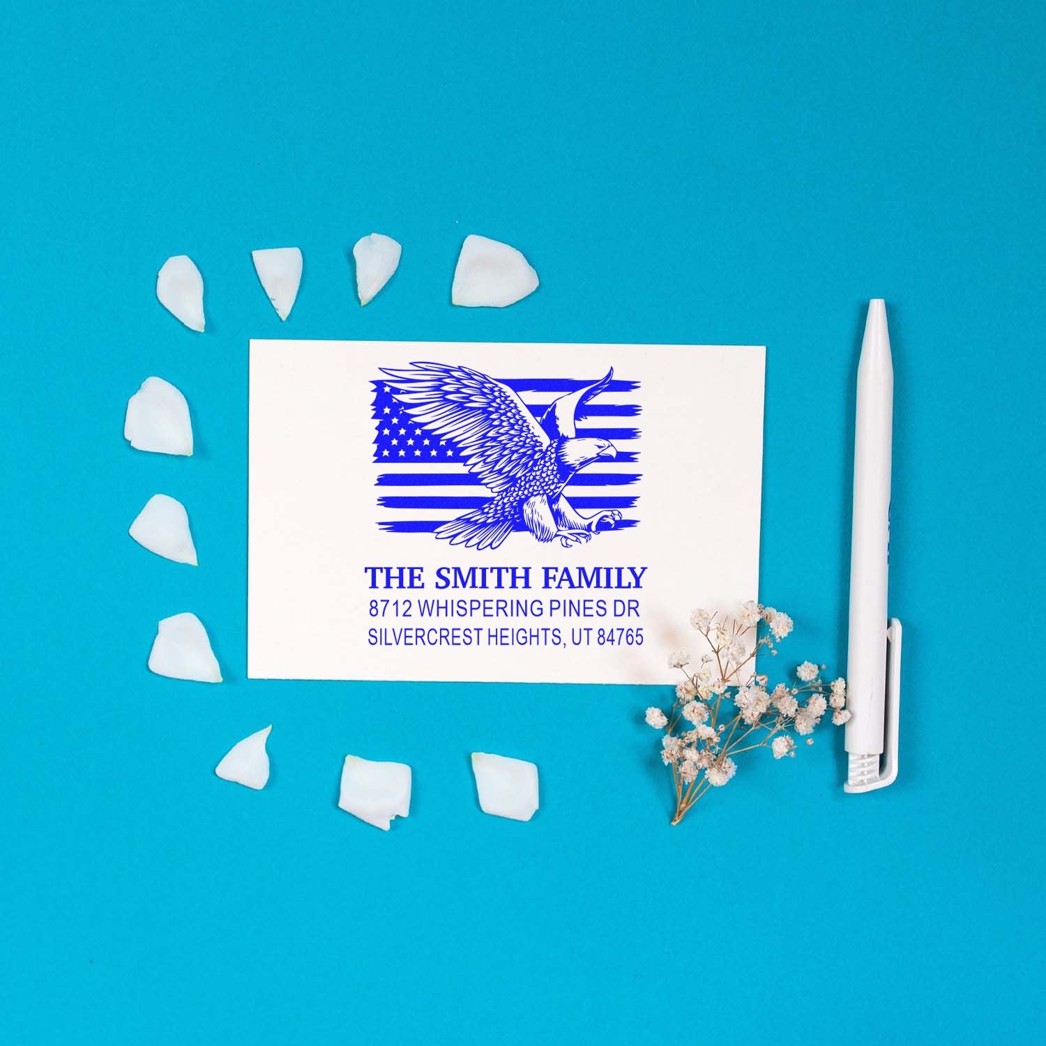 Custom address stamp with eagle and American flag design on blue background, styled with a pen, flower sprigs, and scattered paper petals.