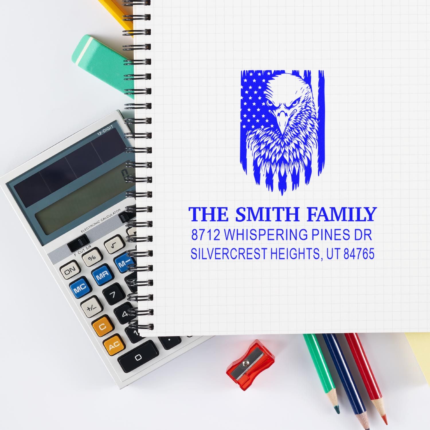 Custom blue family address stamp with eagle design on graph notebook, surrounded by calculator, pencils, eraser and sharpener.
