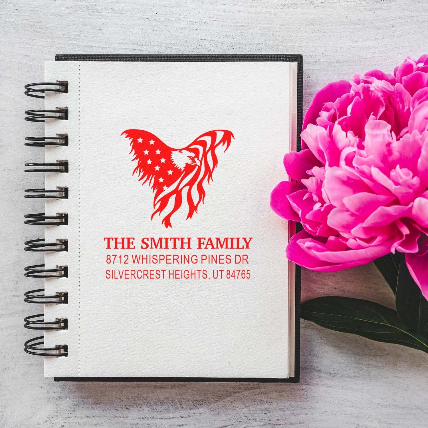 Custom family address stamp with red eagle design on a spiral notebook, styled beside pink peonies on a rustic desk.