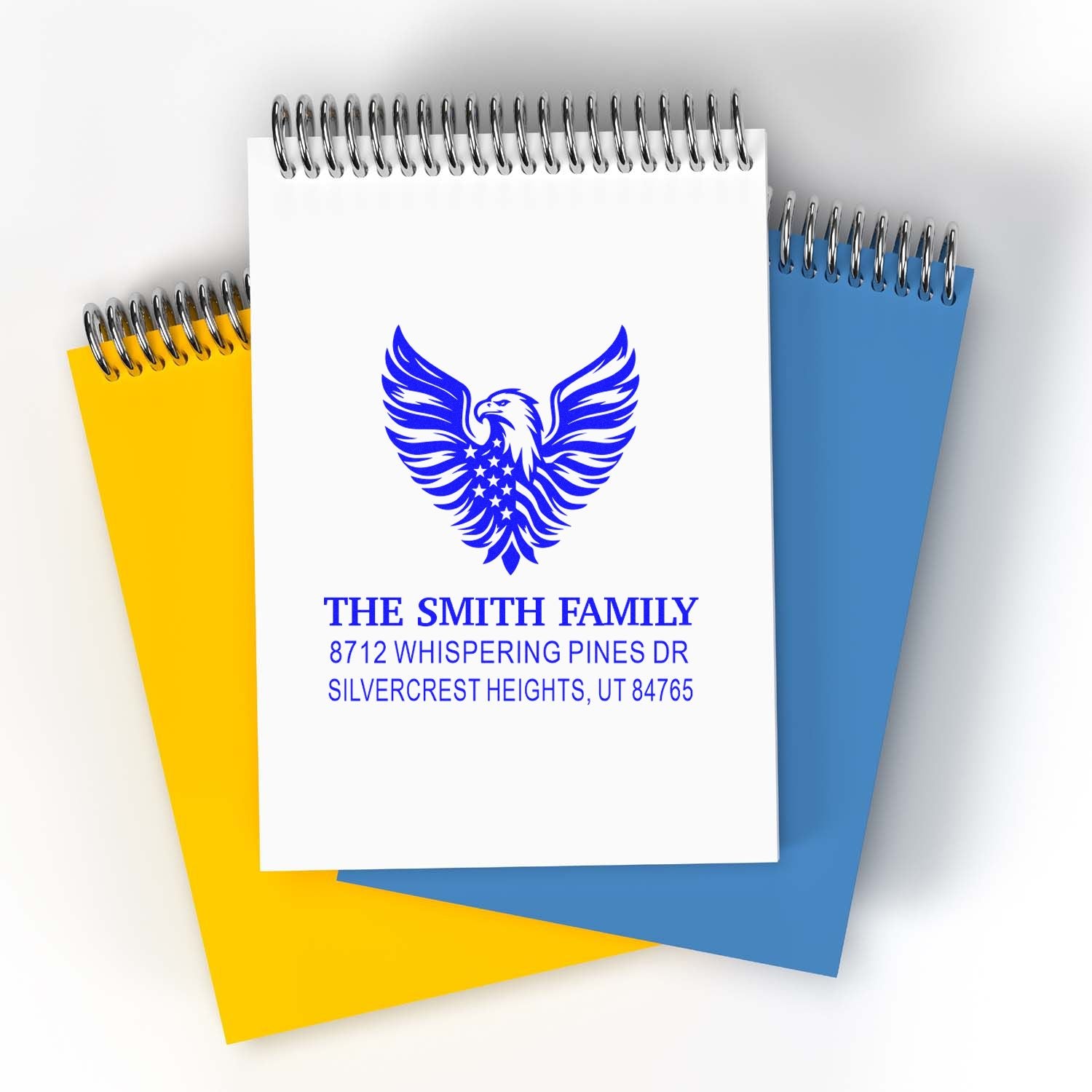 Custom rubber stamp with eagle emblem and family name, shown on spiral notebooks in white, yellow and blue for personalized stationery.