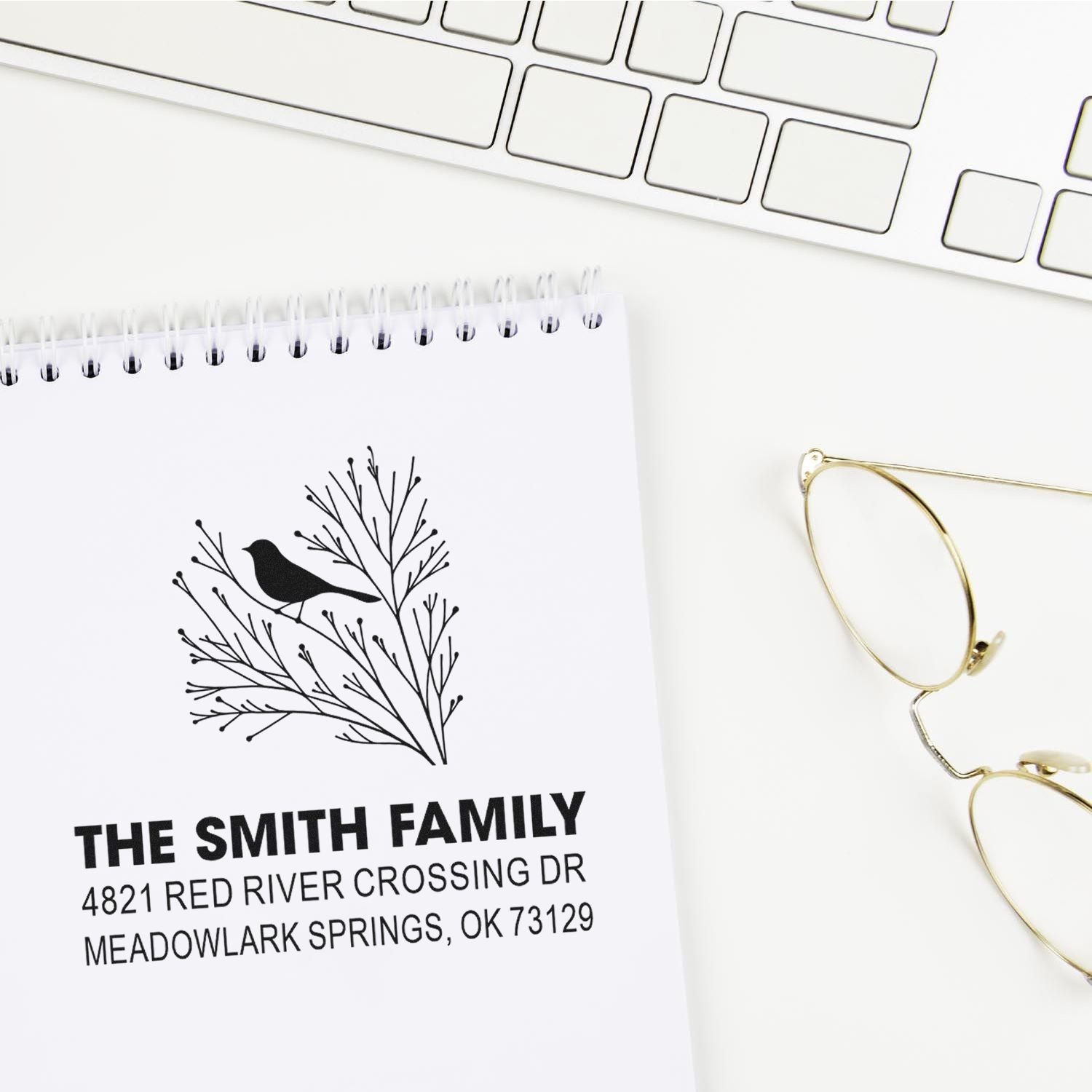 Custom address rubber stamp with bird and branch design, featuring The Smith Family name and address on a notebook beside a keyboard and glasses.