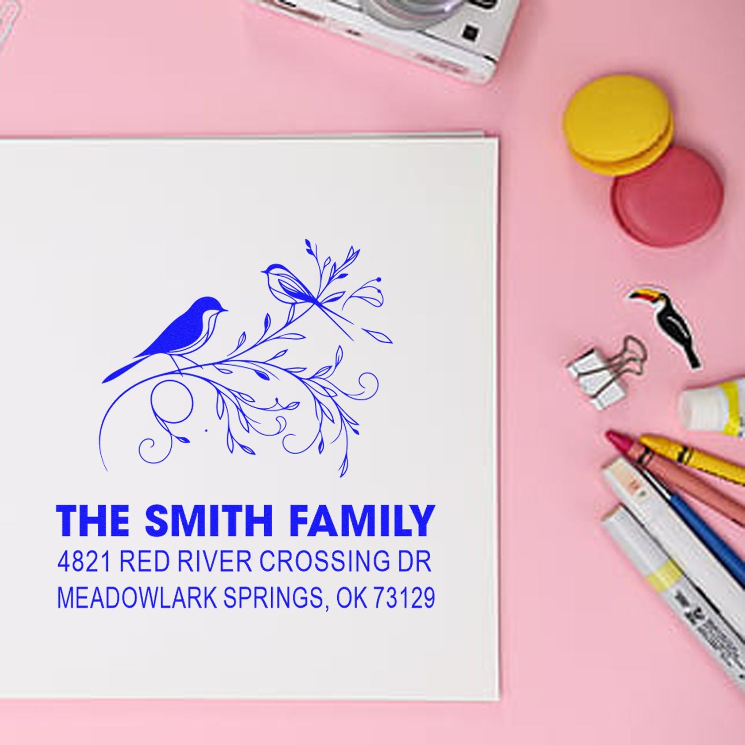 Custom self-inking rubber address stamp with bird design, family name and return address in blue, shown with stamping supplies on pink background.