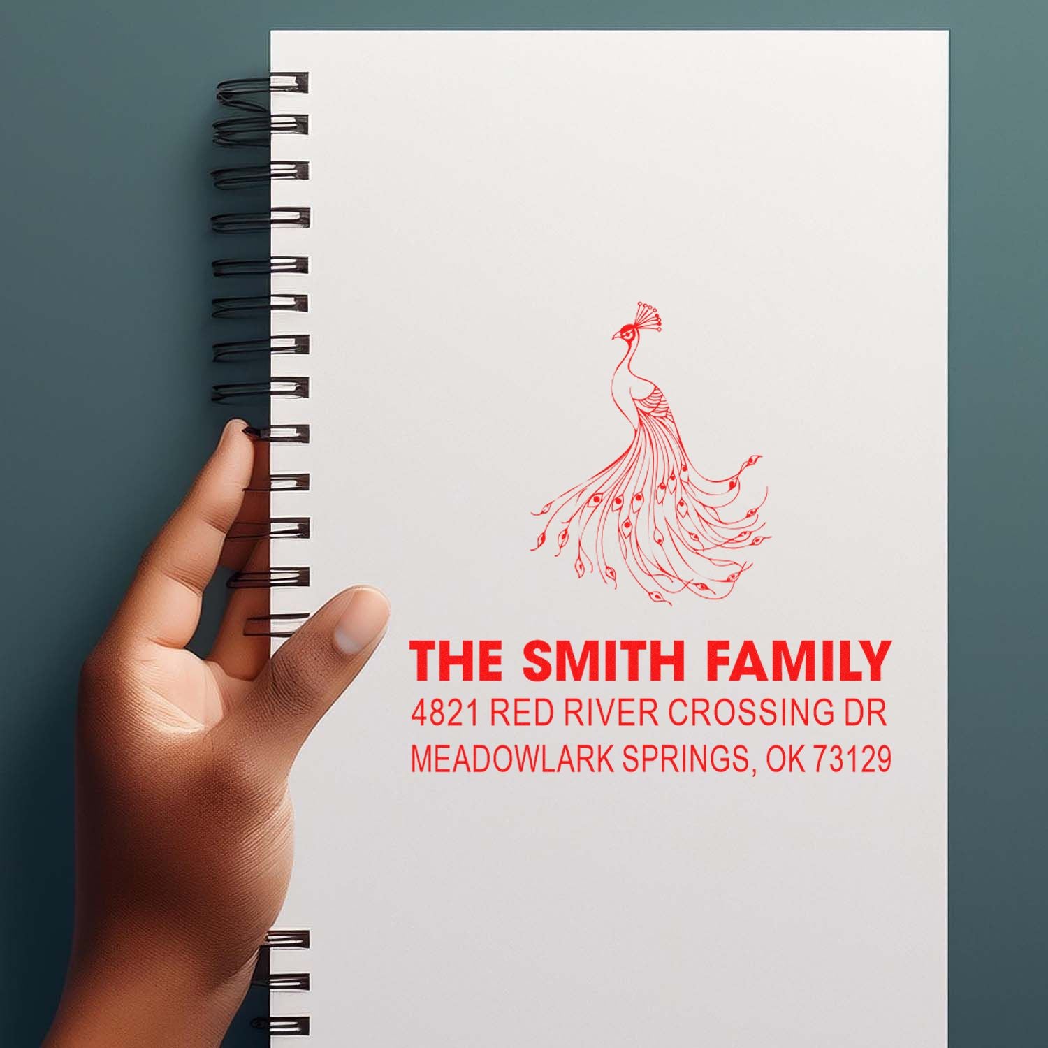 Personalized rubber stamp with red peacock design and family return address, stamped on a white spiral notebook for custom stationery and mailing