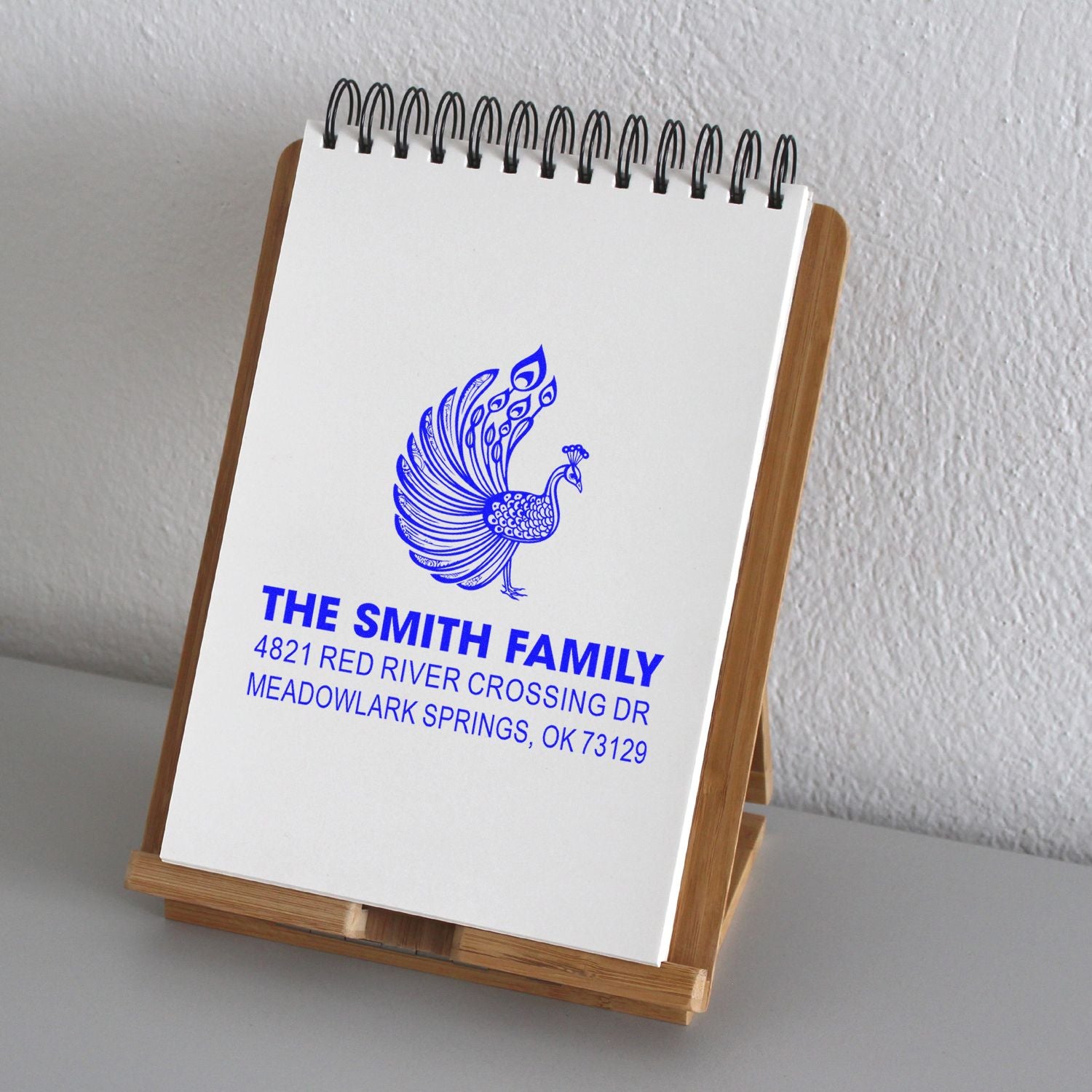 Custom family return address rubber stamp with peacock design, blue ink impression, and personalized name and address on notepad stand