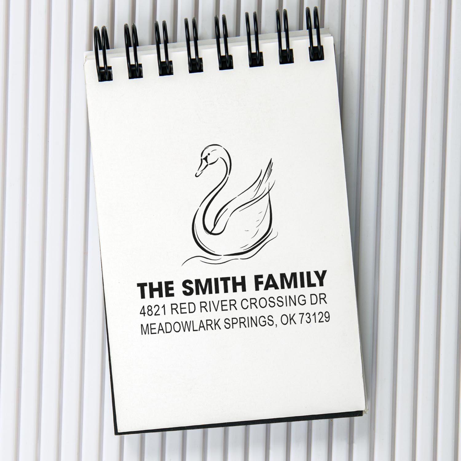 Custom rubber stamp with swan design, family name, and return address details on a spiral notepad for personalized mailing and stationery use.