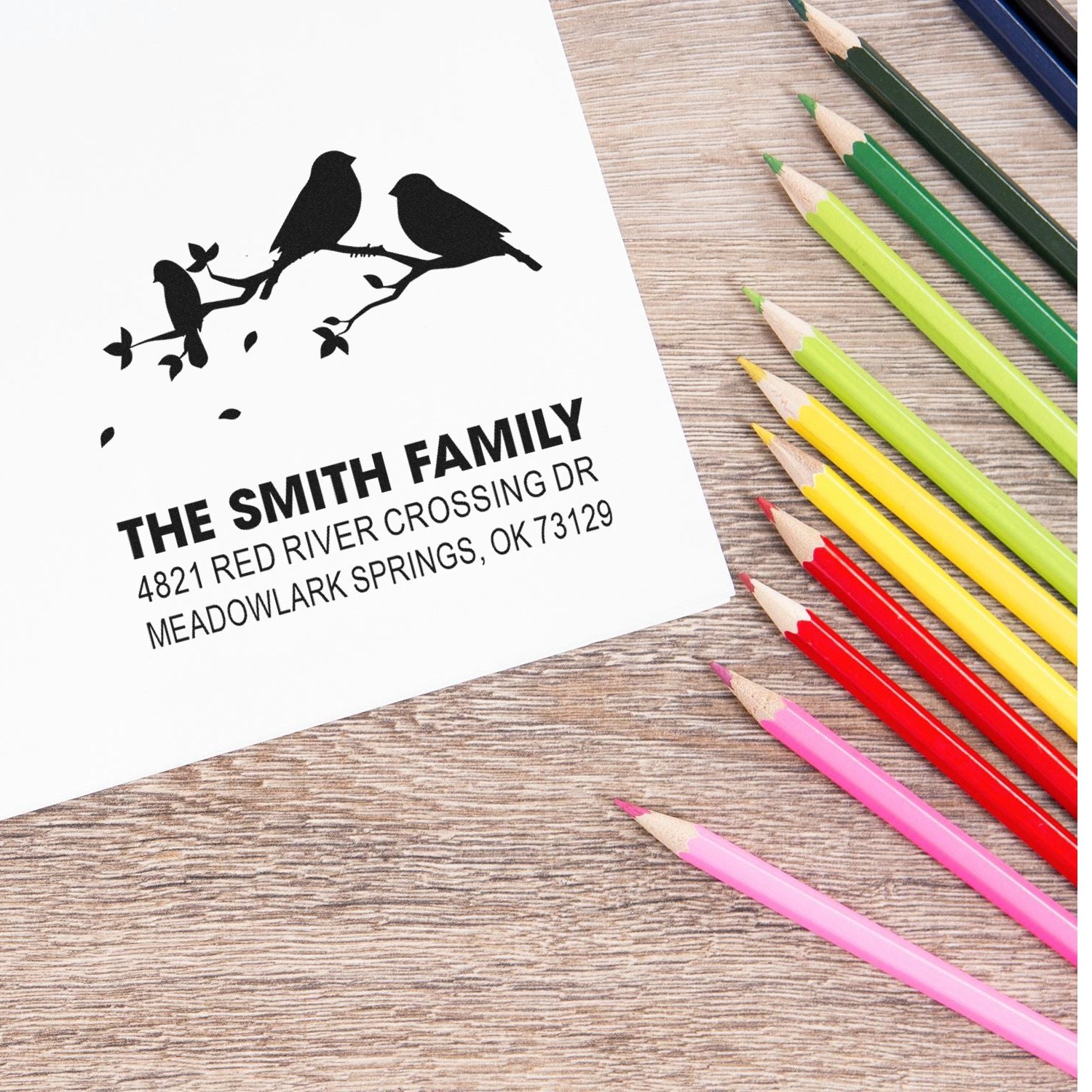Personalized return address rubber stamp with bird branch design and The Smith Family address on white paper, shown beside colorful pencils.