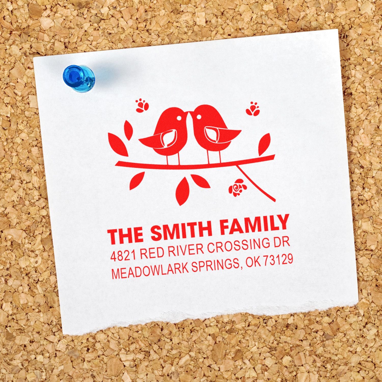 Custom rubber address stamp with red bird design, featuring family name and mailing address on white paper pinned to a corkboard background.