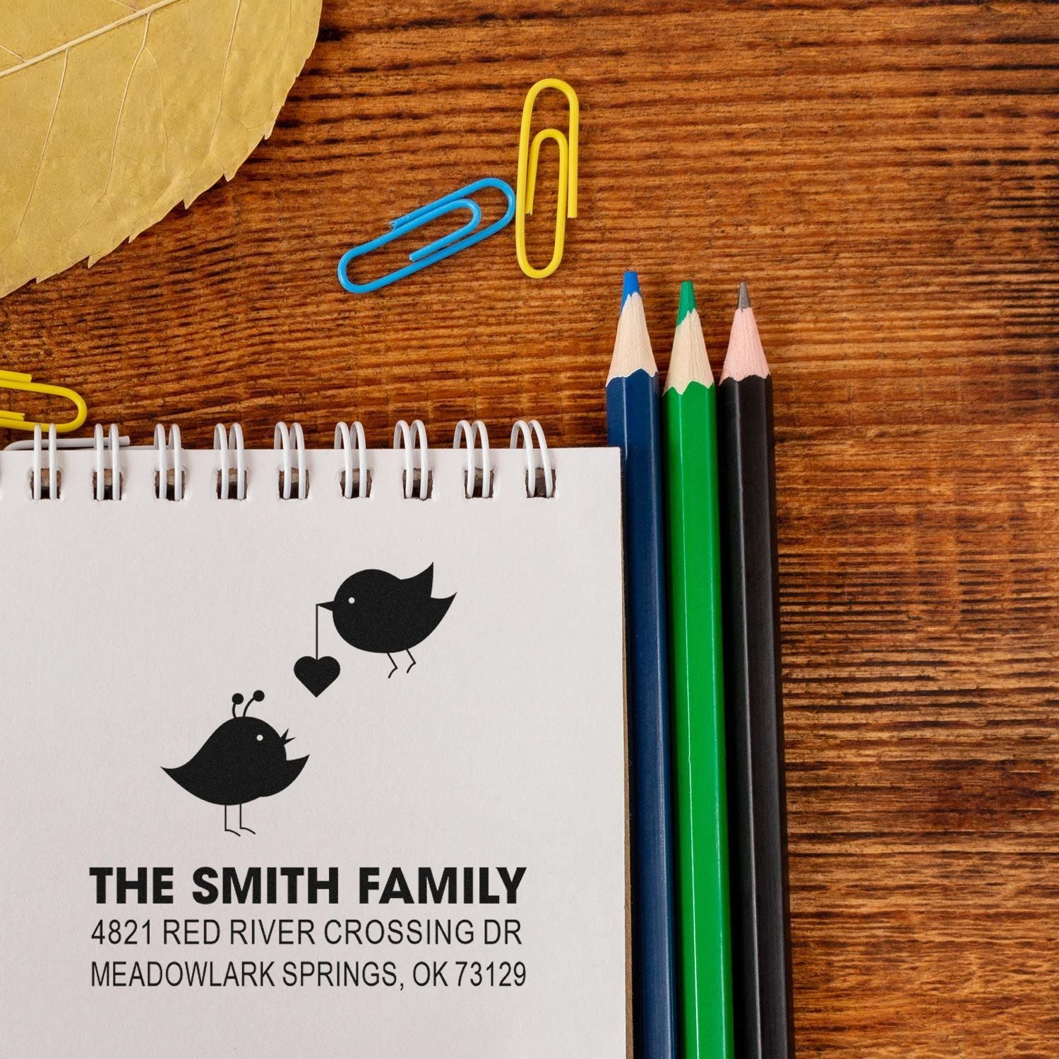 Custom family address rubber stamp impression with bird design on spiral notebook, surrounded by colored pencils and paper clips on wood desk.