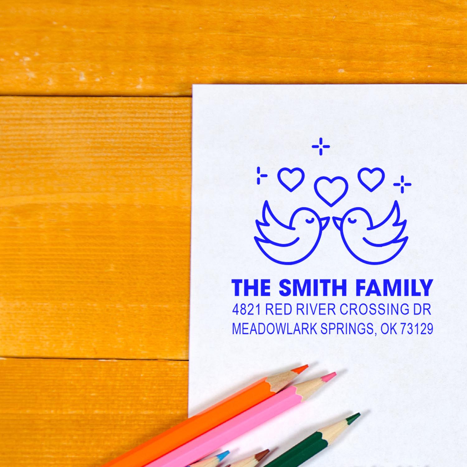 Personalized family return address rubber stamp with birds and hearts, blue imprint on white paper beside colored pencils on a wooden desk for mailings.