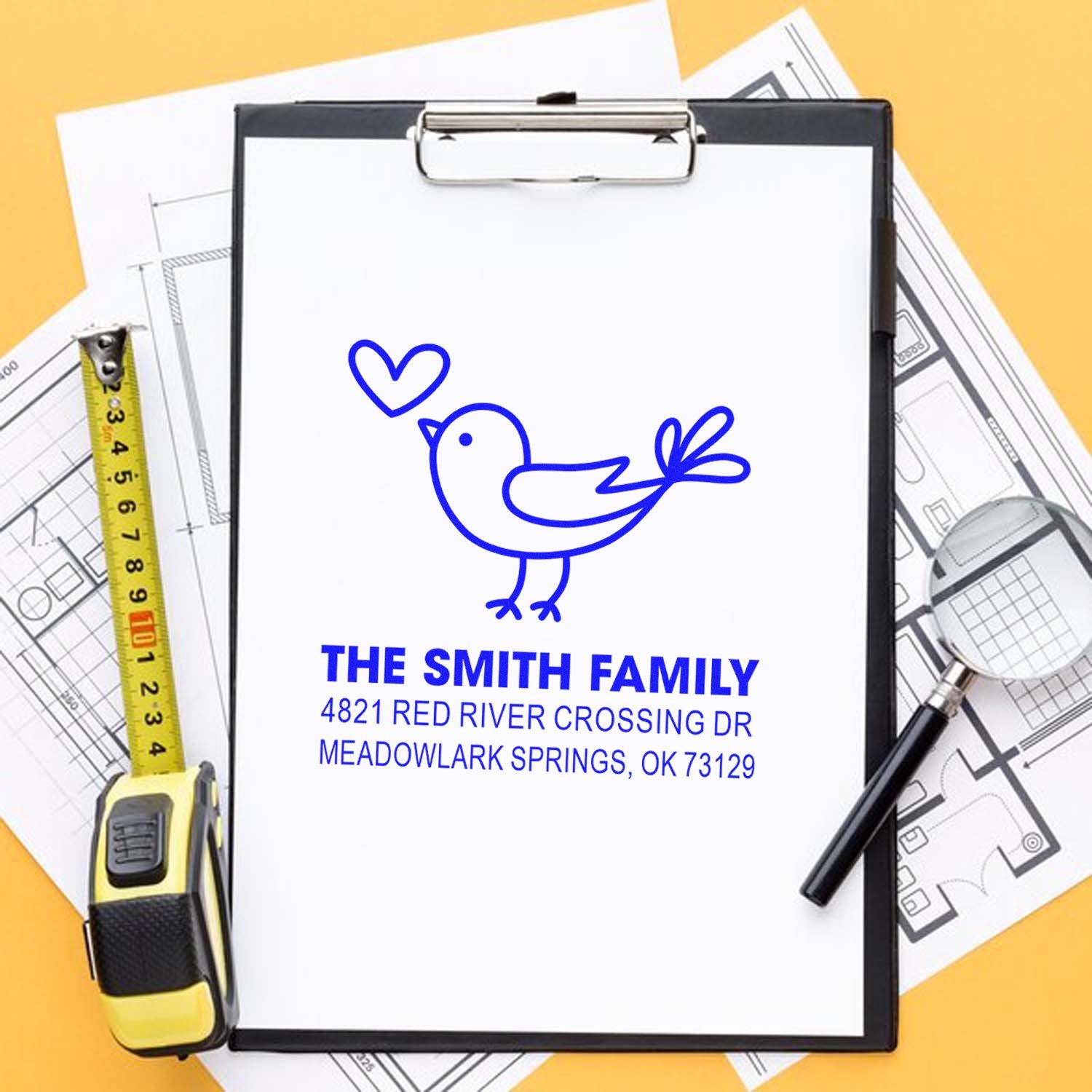 Custom return address rubber stamp with bird and heart design, personalized family name and address in blue ink on clipboard with measuring tape and plans