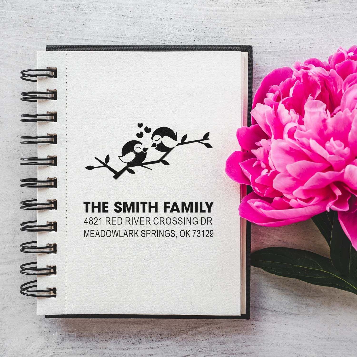 Personalized return address rubber stamp with bird design, stamped on a notebook for The Smith Family, beside bright pink peonies and a mailing address.