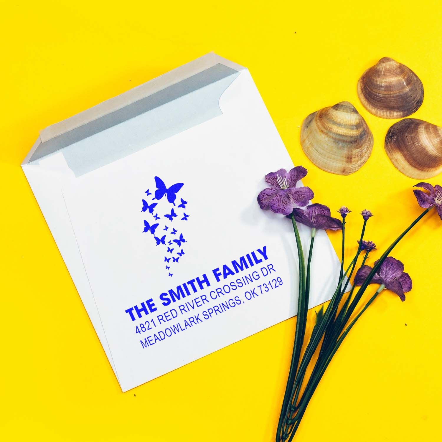 Custom return address rubber stamp on white envelope with butterfly design, blue family name and address, styled with seashells and purple flowers.