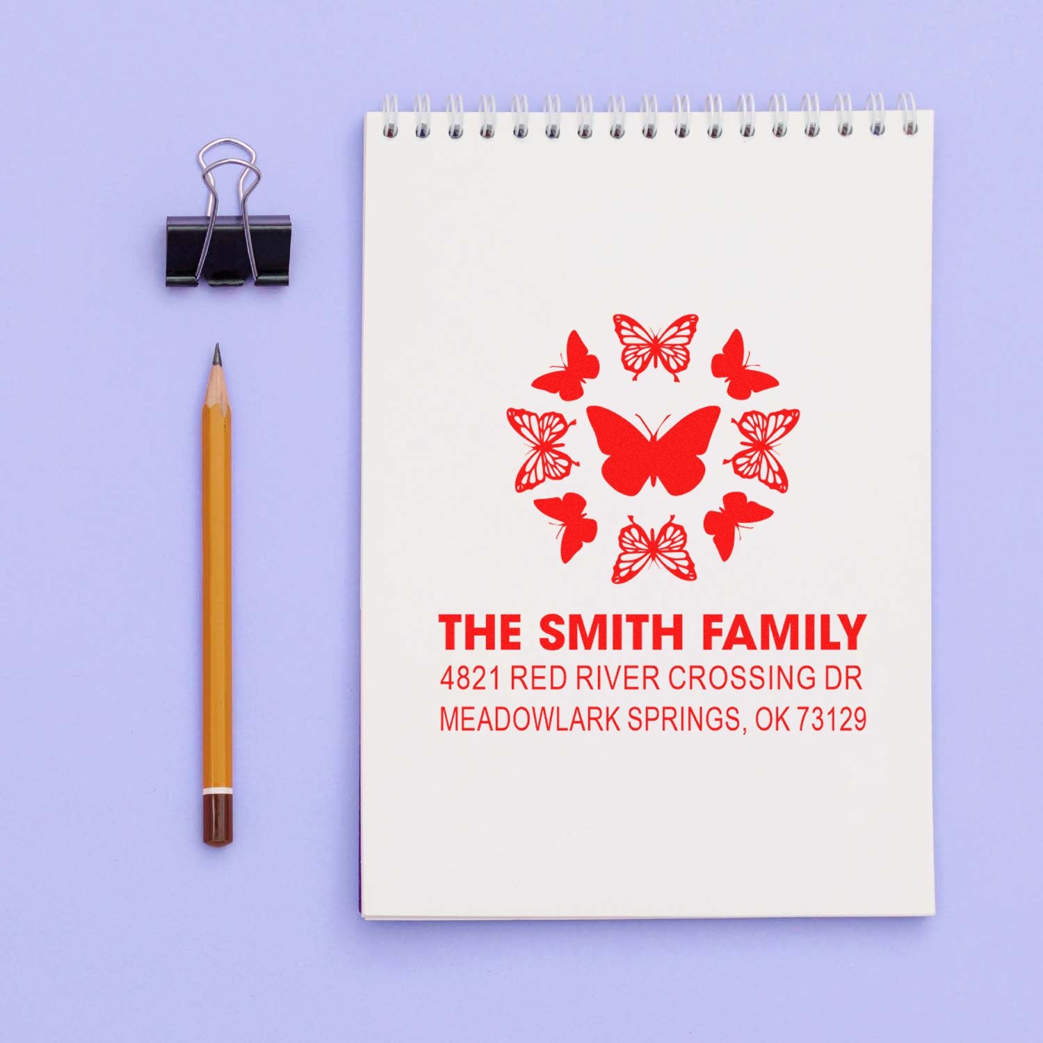 Personalized red butterfly family address rubber stamp with custom name and mailing address, displayed on a spiral notebook with pencil and binder clip.